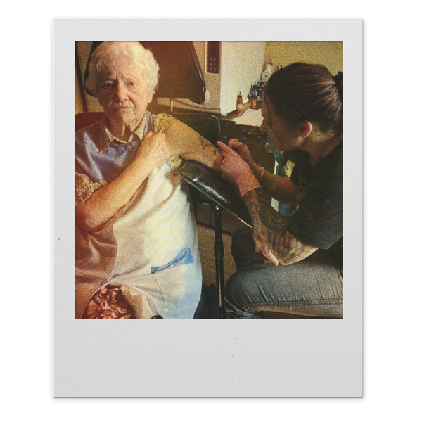 An elderly woman with white hair sits in a wheelchair while a tattoo artist tattoos her arm in a room.