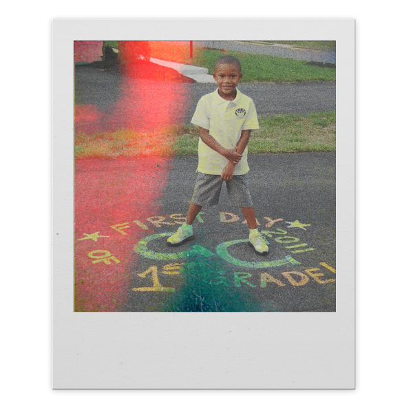 A young boy in a yellow school uniform standing on a sidewalk with colorful chalk writing that reads "First Day of 1st Grade" around him. The boy is smiling and has short hair.