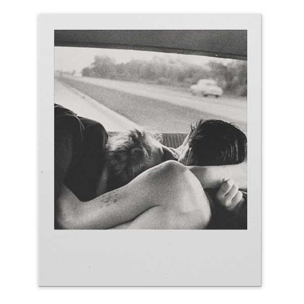 Two people are sleeping in the back seat of a car, with one hugging the other, during a road trip.