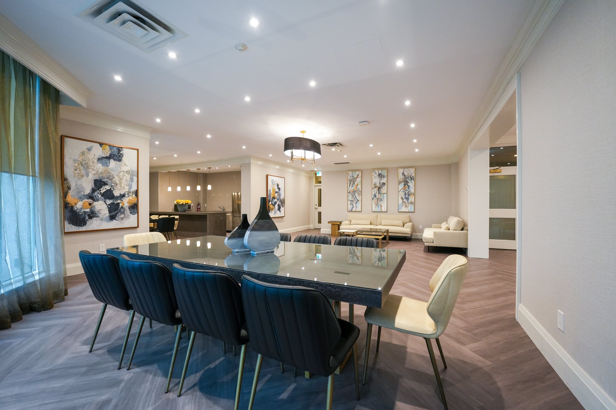 Modern dining and living room area with a large glass table, black and beige chairs, abstract paintings, and light-colored couches.