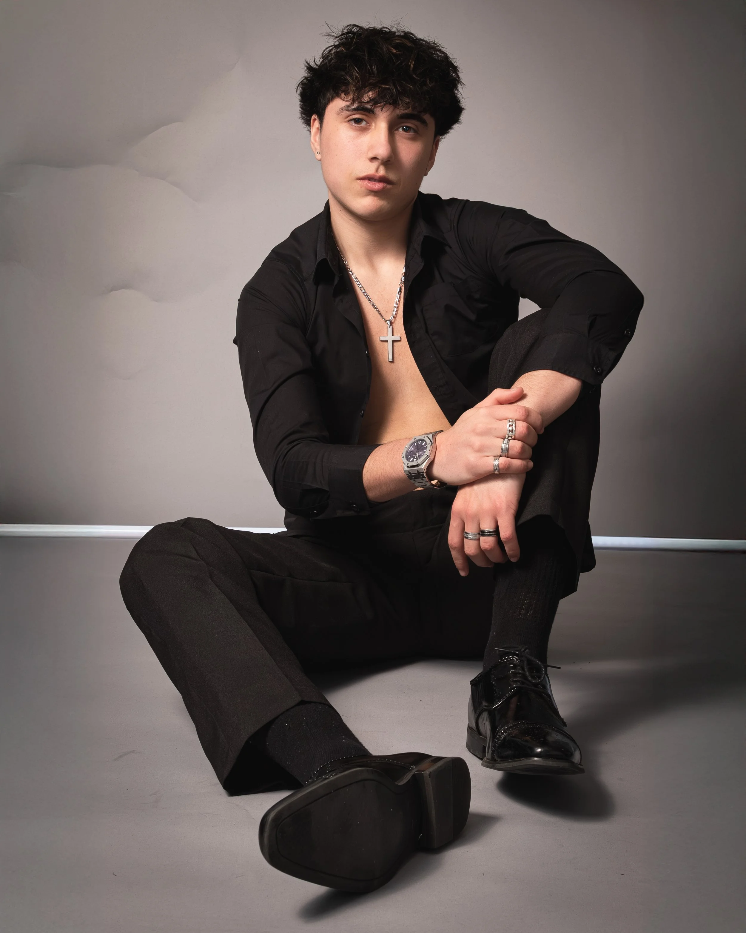 Young man with dark hair sitting on the floor, wearing a black shirt, black pants, black shiny shoes, a silver watch, multiple rings, and a silver cross necklace. The shirt is partly unbuttoned, revealing his chest. The background is plain with a gra