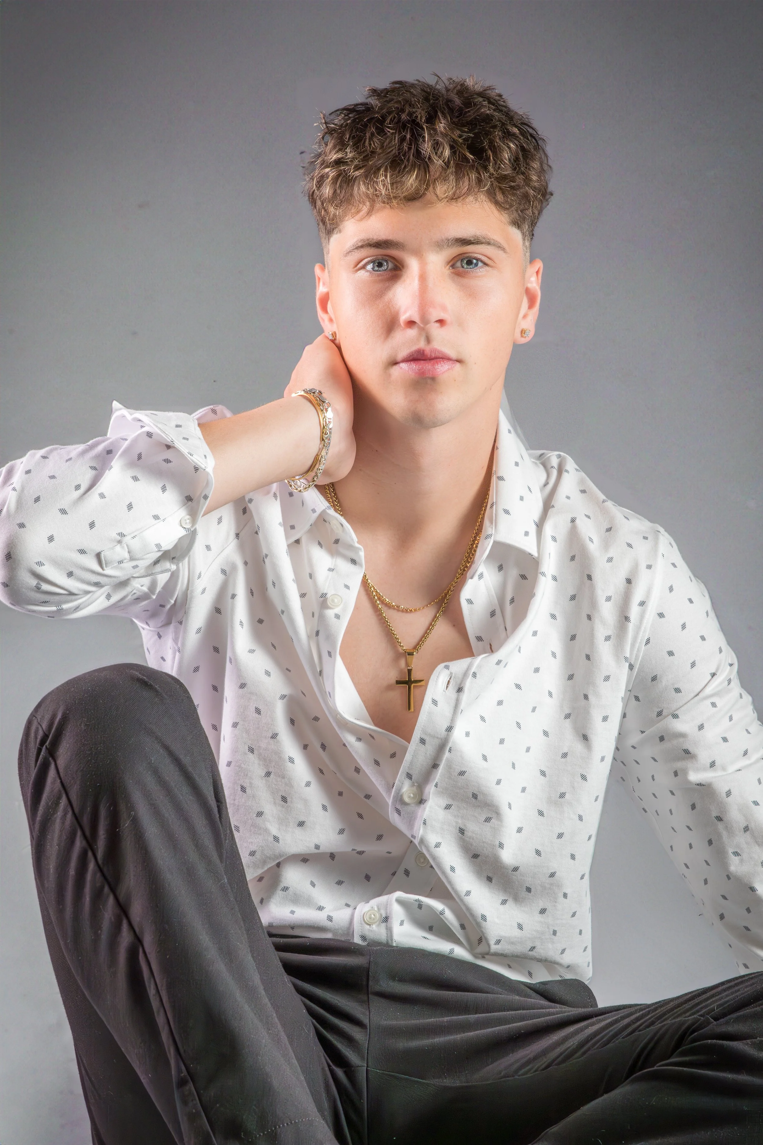 Young man with short, curly brown hair and light skin, sitting against a neutral gray background, wearing a white shirt with small black dots, black pants, gold jewelry including a cross necklace and earrings, with a serious expression, resting his r
