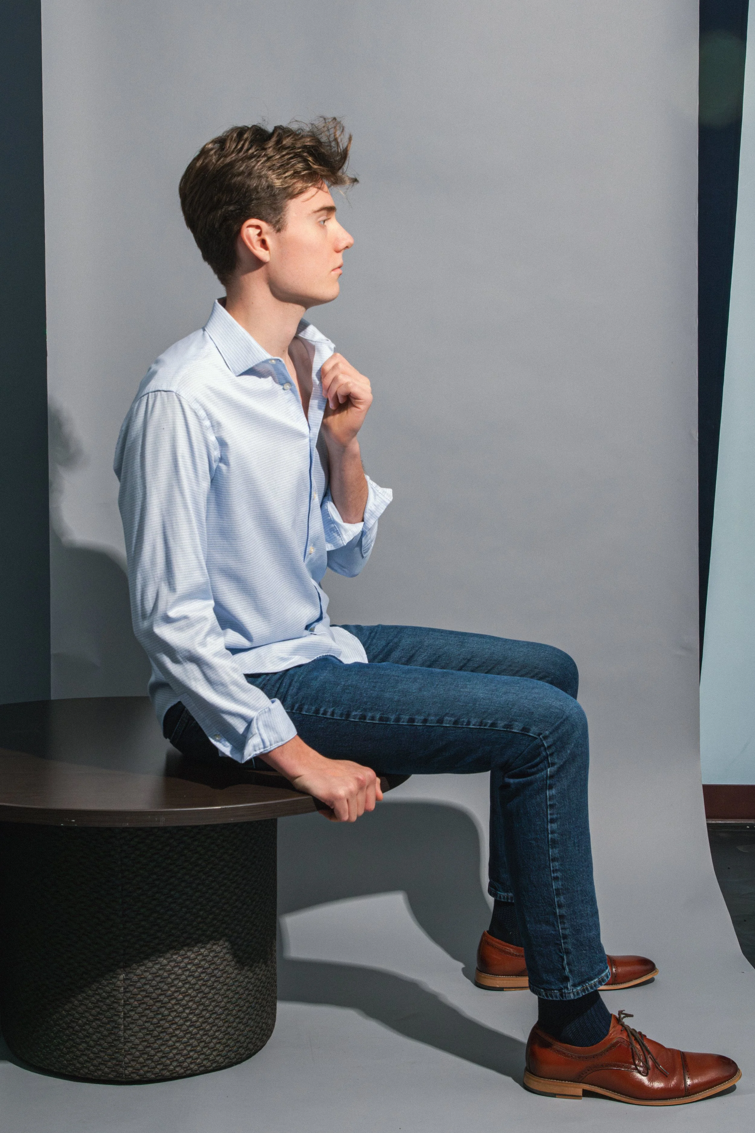 Young man with brown hair in a side profile, wearing a light blue dress shirt, dark jeans, and brown dress shoes, sitting on a dark textured cylinder with a light gray background.