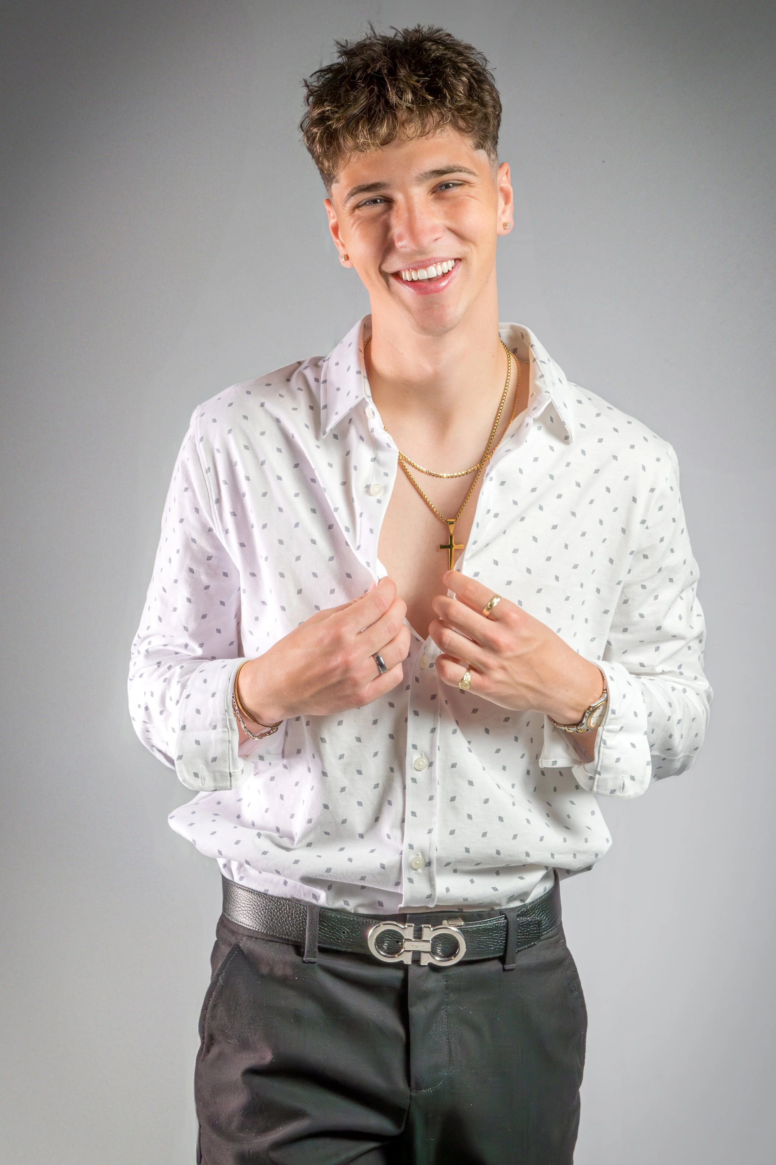 A young man smiling, wearing a white shirt with small black dots, gray pants, and accessorized with jewelry including necklaces, rings, earrings, a watch, and a belt with a distinctive logo, standing against a plain gray background.