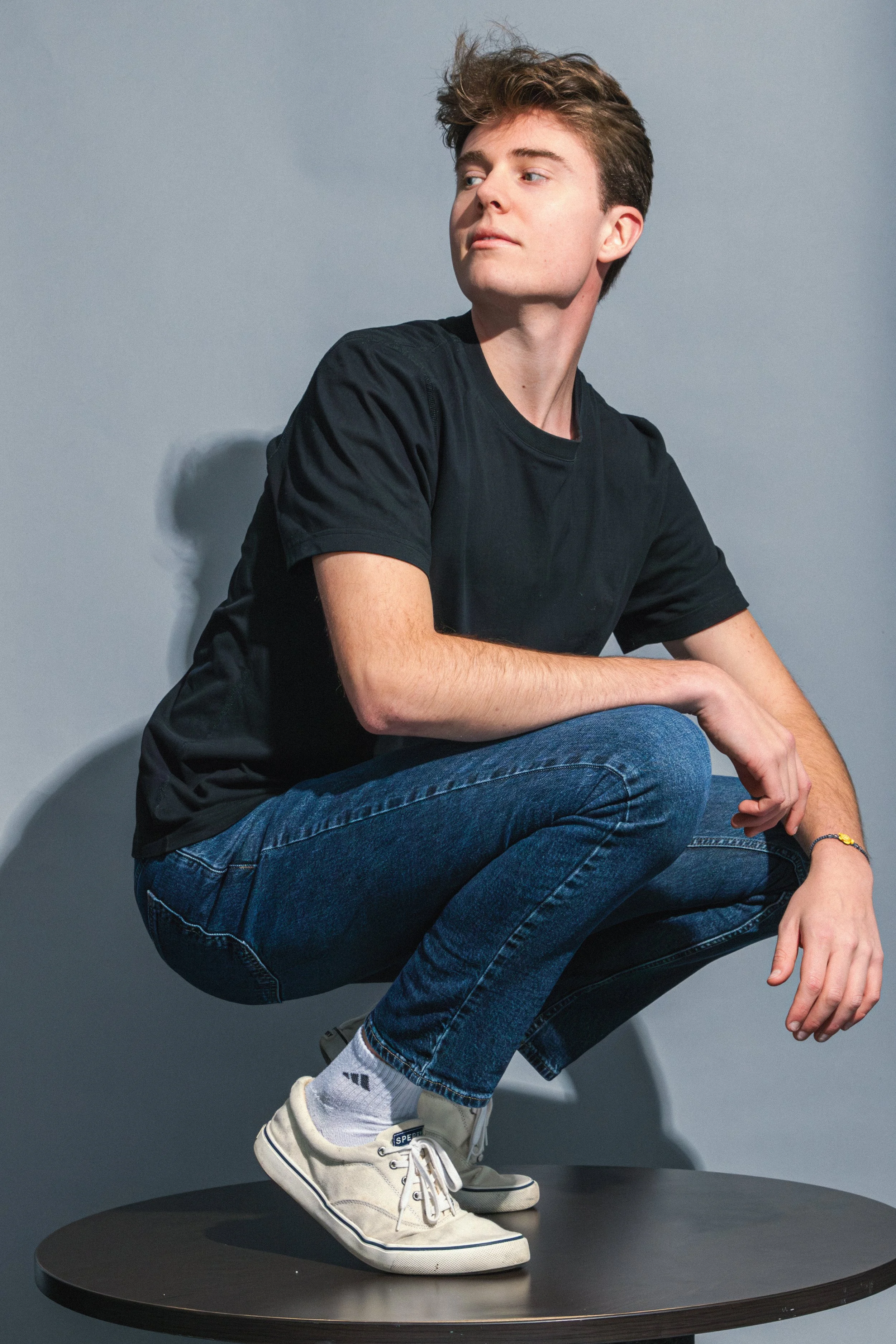 A young man squatting on a round black table against a gray background, with a casual pose, wearing a black T-shirt, blue jeans, white sneakers, and white socks.