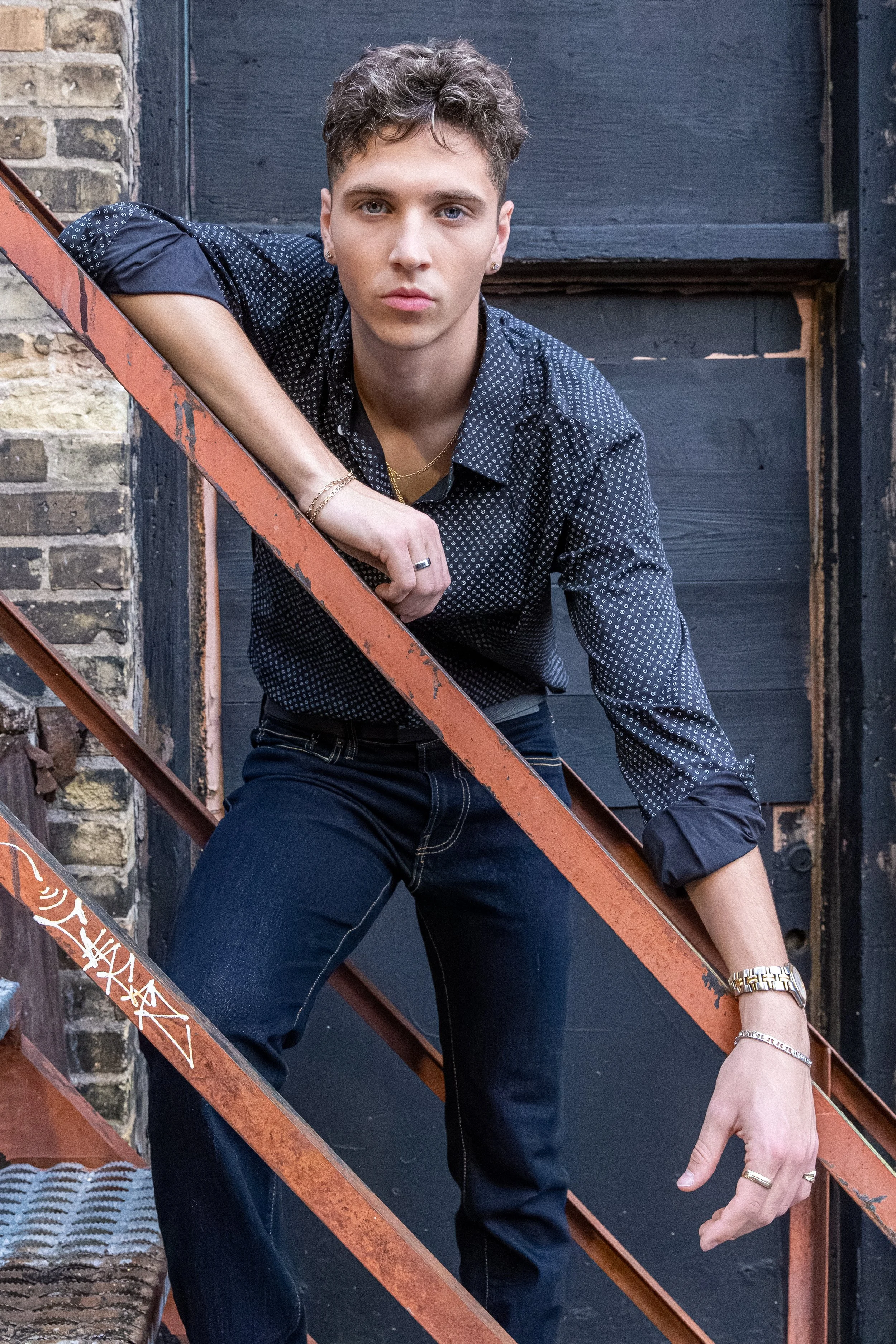 A young man with curly hair leaning on a rusty orange metal staircase, looking at the camera, wearing a dark patterned shirt, dark jeans, and jewelry, with a brick wall and black wooden door behind him.