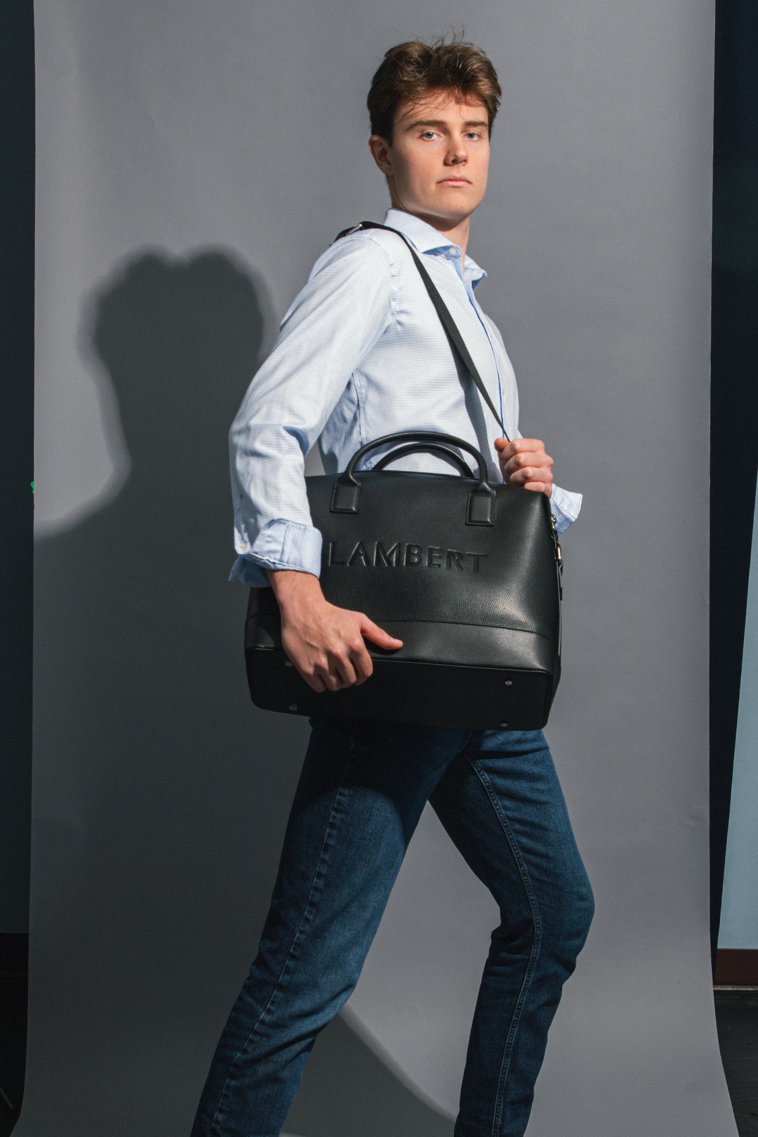 A young man with brown hair dressed in a light blue shirt and blue jeans, holding a black leather bag with the word 'LAMBERT' embossed on it, standing against a gray background.