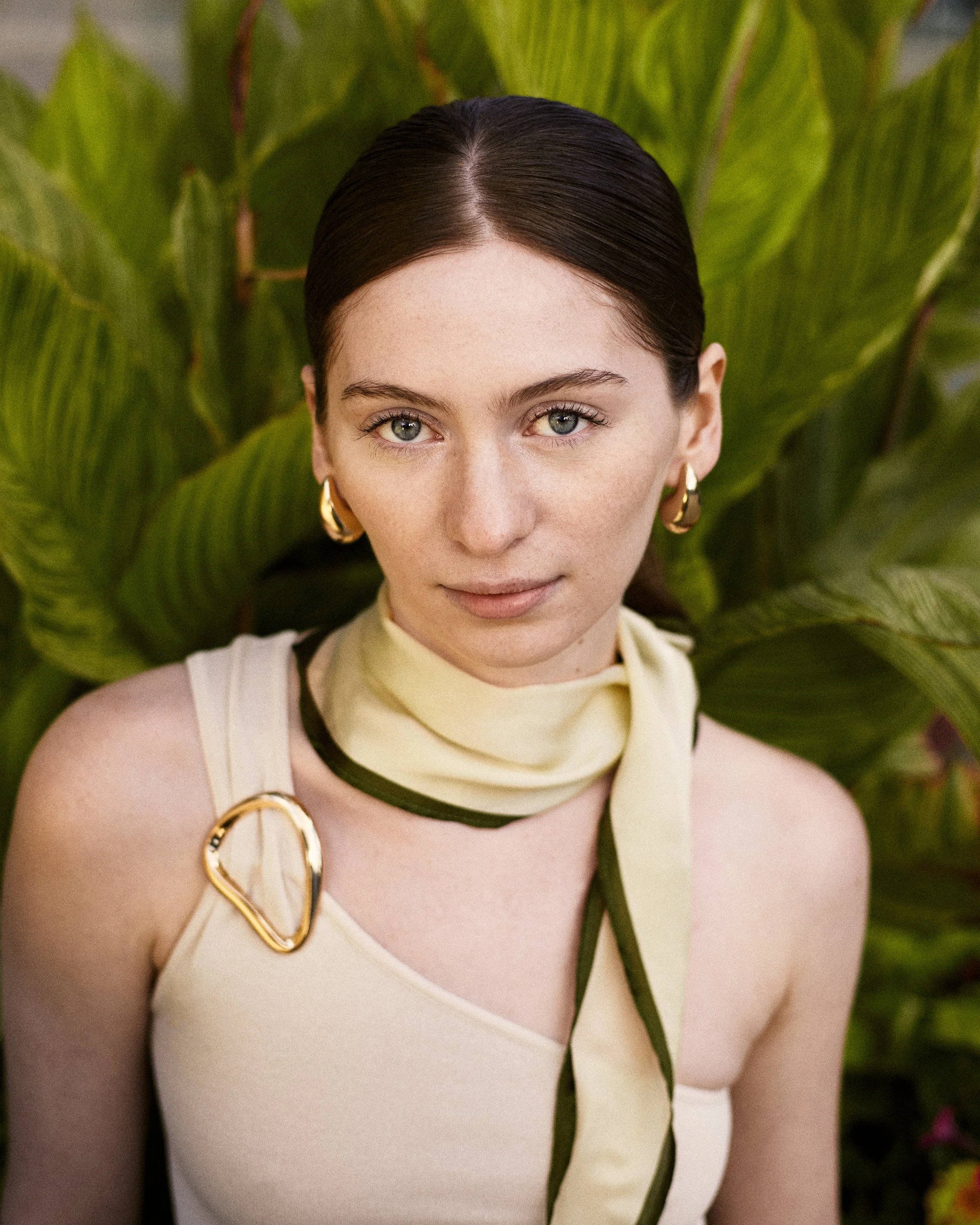 A woman with dark hair, blue eyes, and gold hoop earrings stands in front of green tropical leaves. She wears a beige top with a decorative gold pin and a light-colored silk scarf around her neck with green accents.