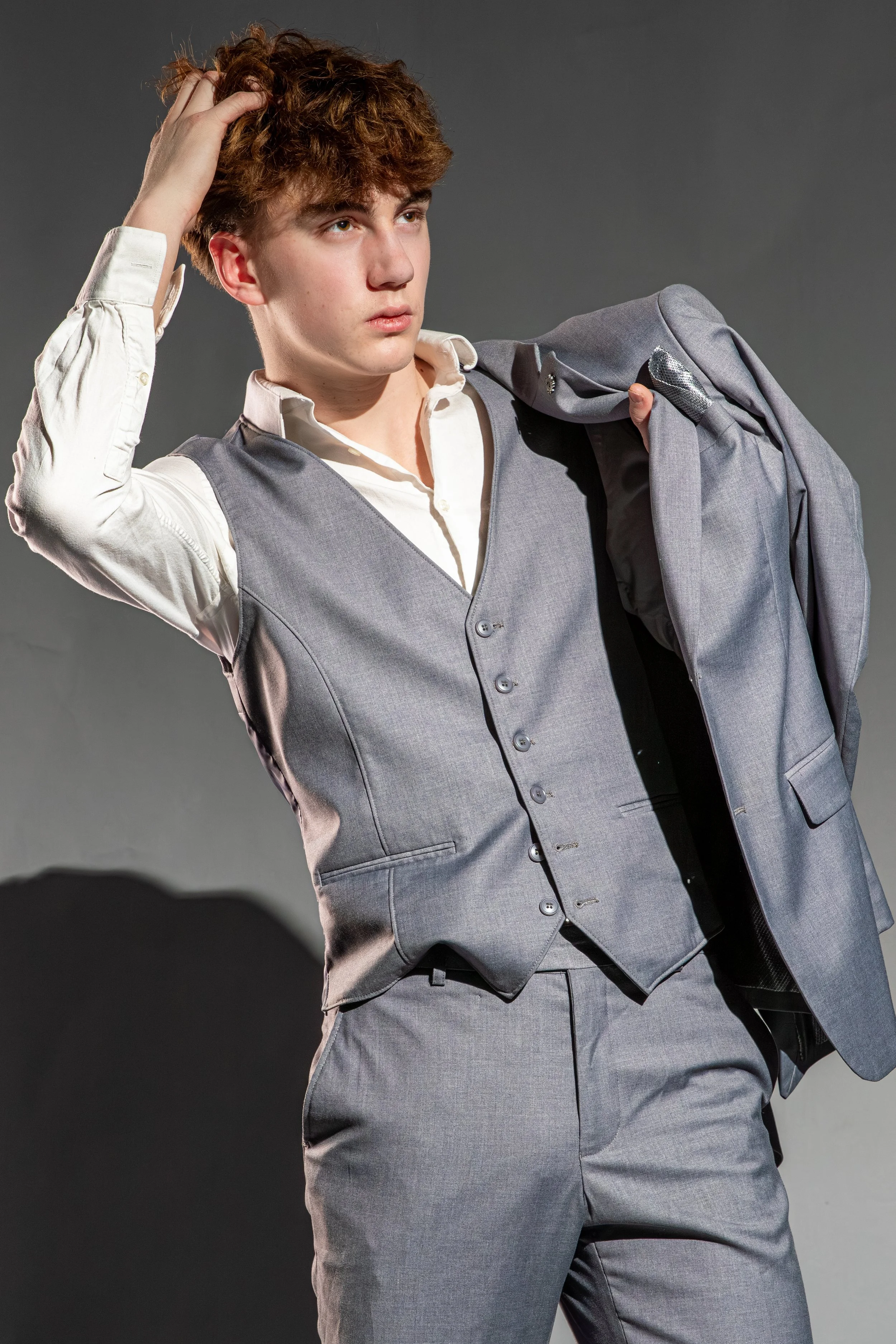 A young man dressed in a gray vest, white shirt, and matching gray pants, holding a gray suit jacket over his shoulder, with one hand in his hair, looking serious against a gray background.