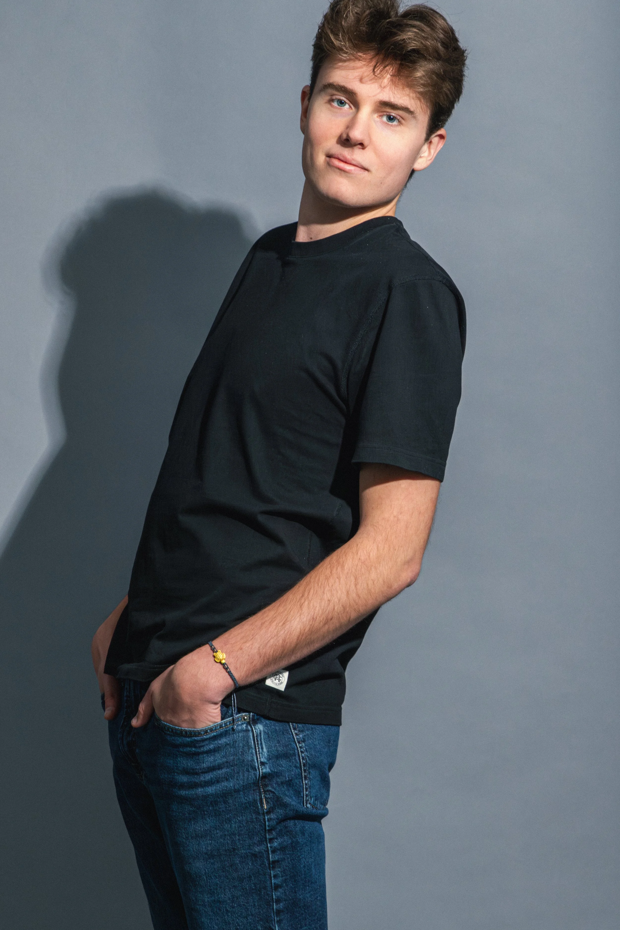 A young man with light brown hair and blue eyes, wearing a black T-shirt and jeans, standing with his hands in his pockets against a plain gray background, casting a shadow.