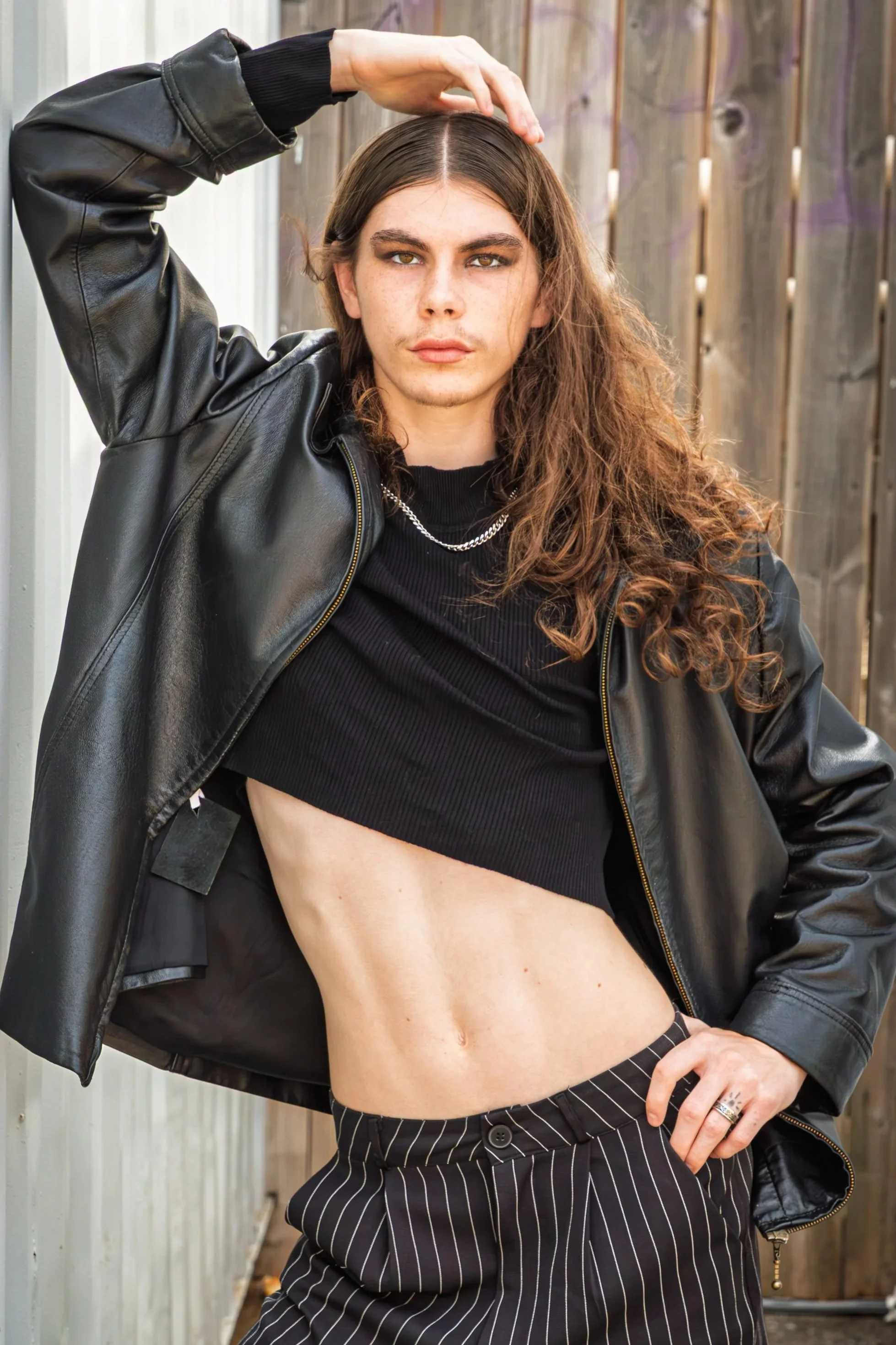 A young woman with long curly brown hair, wearing a black leather jacket, black crop top, and pinstriped pants, posing outdoors with a wooden fence in the background.