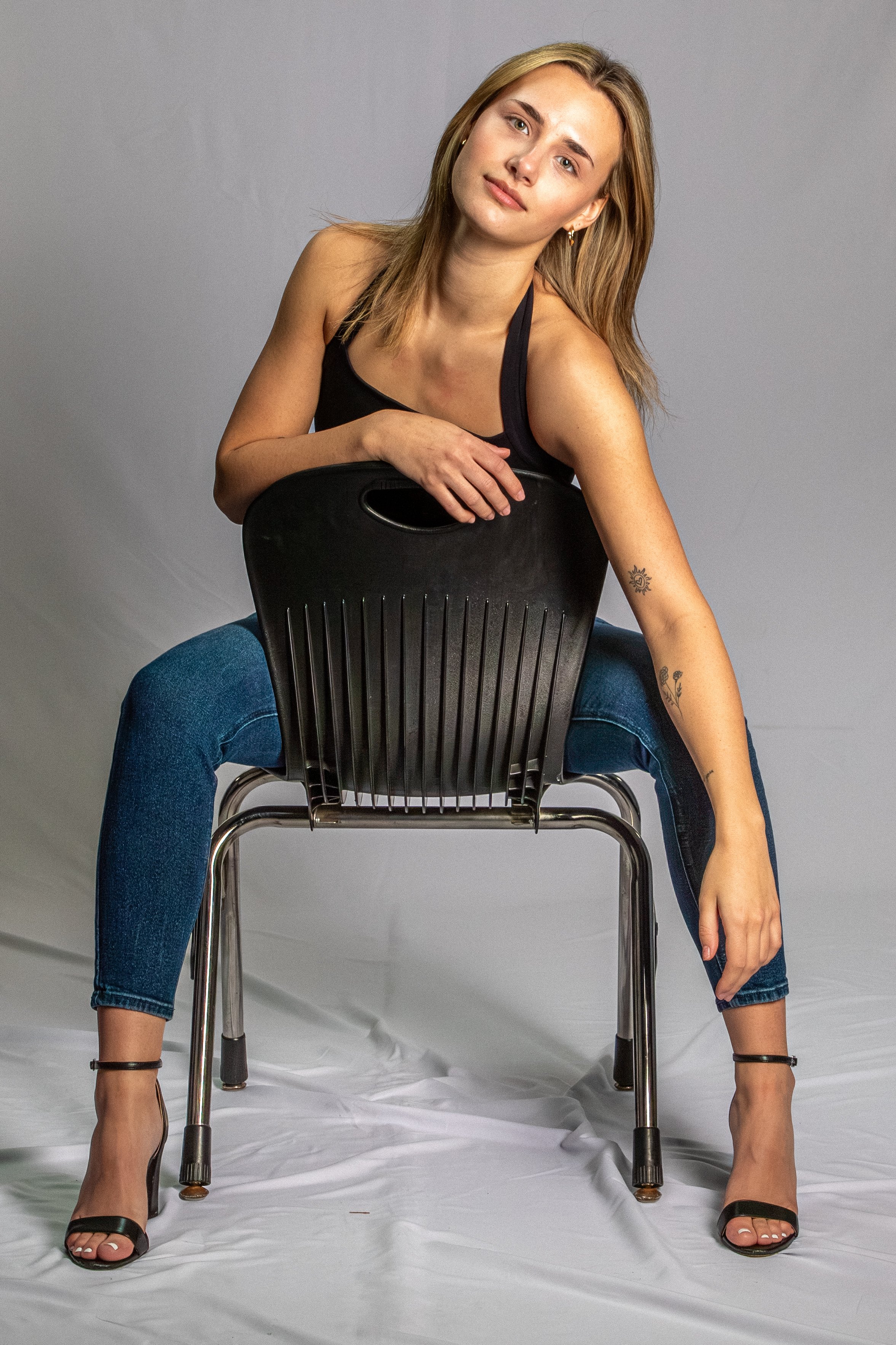 A young woman with blonde hair, wearing a black tank top and jeans, is seated on a black plastic chair with her legs spread apart, her feet in black high-heeled sandals, and her arms resting on the back of the chair. She has tattoos on her right arm 