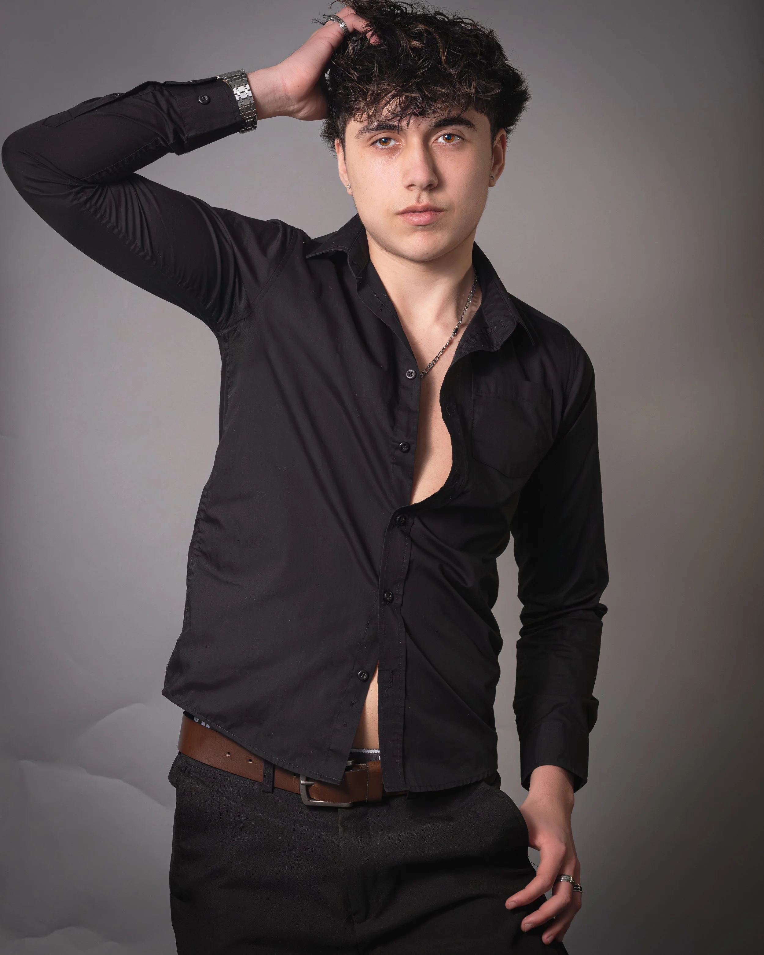 Young man with curly dark hair wearing a black button-up shirt, unbuttoned at the top, posing with one hand in his pocket and the other hand on his head, against a gray background.