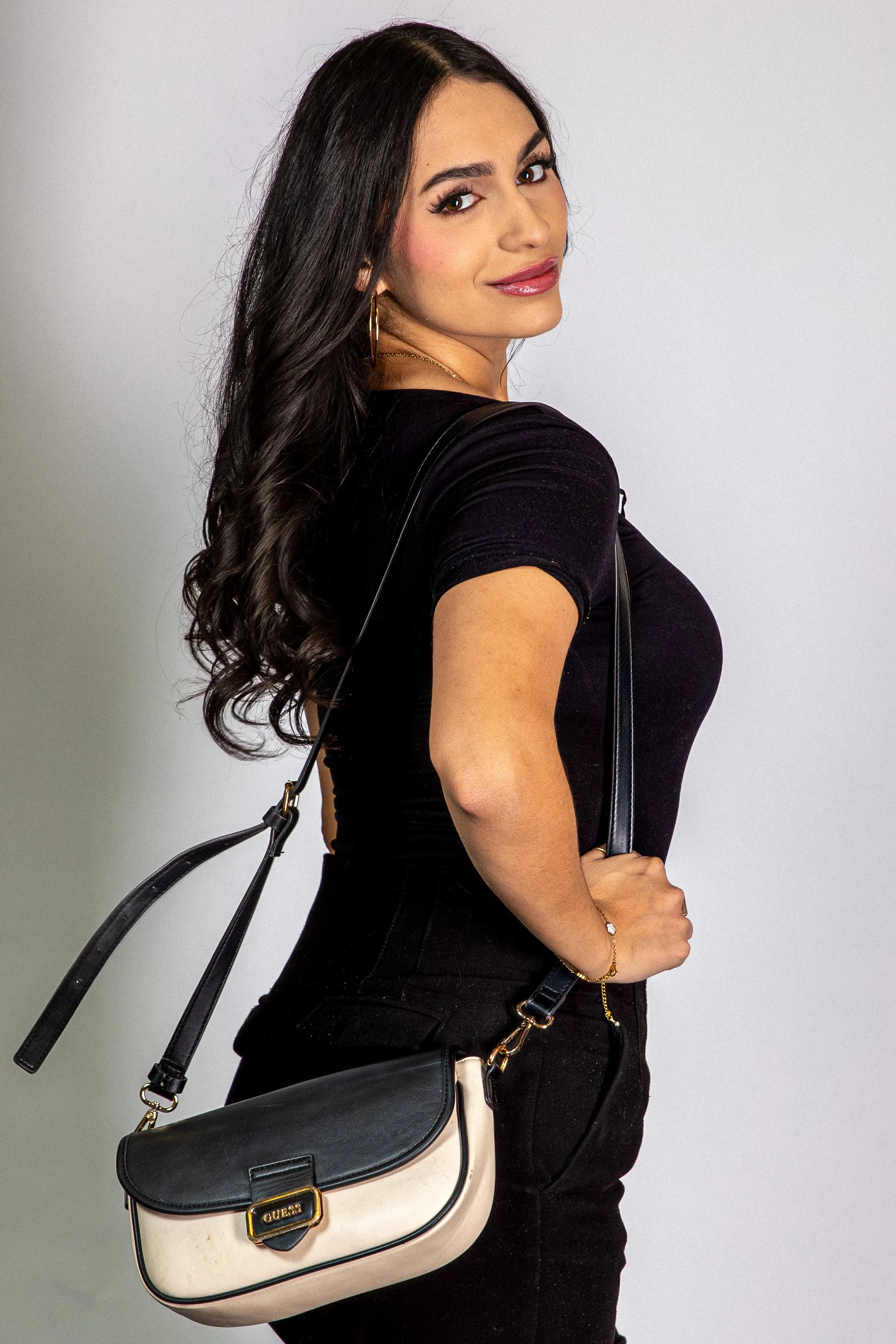Young woman with long dark hair, wearing a black t-shirt, black pants, and gold jewelry, posing with a stylish black and beige Guess purse.