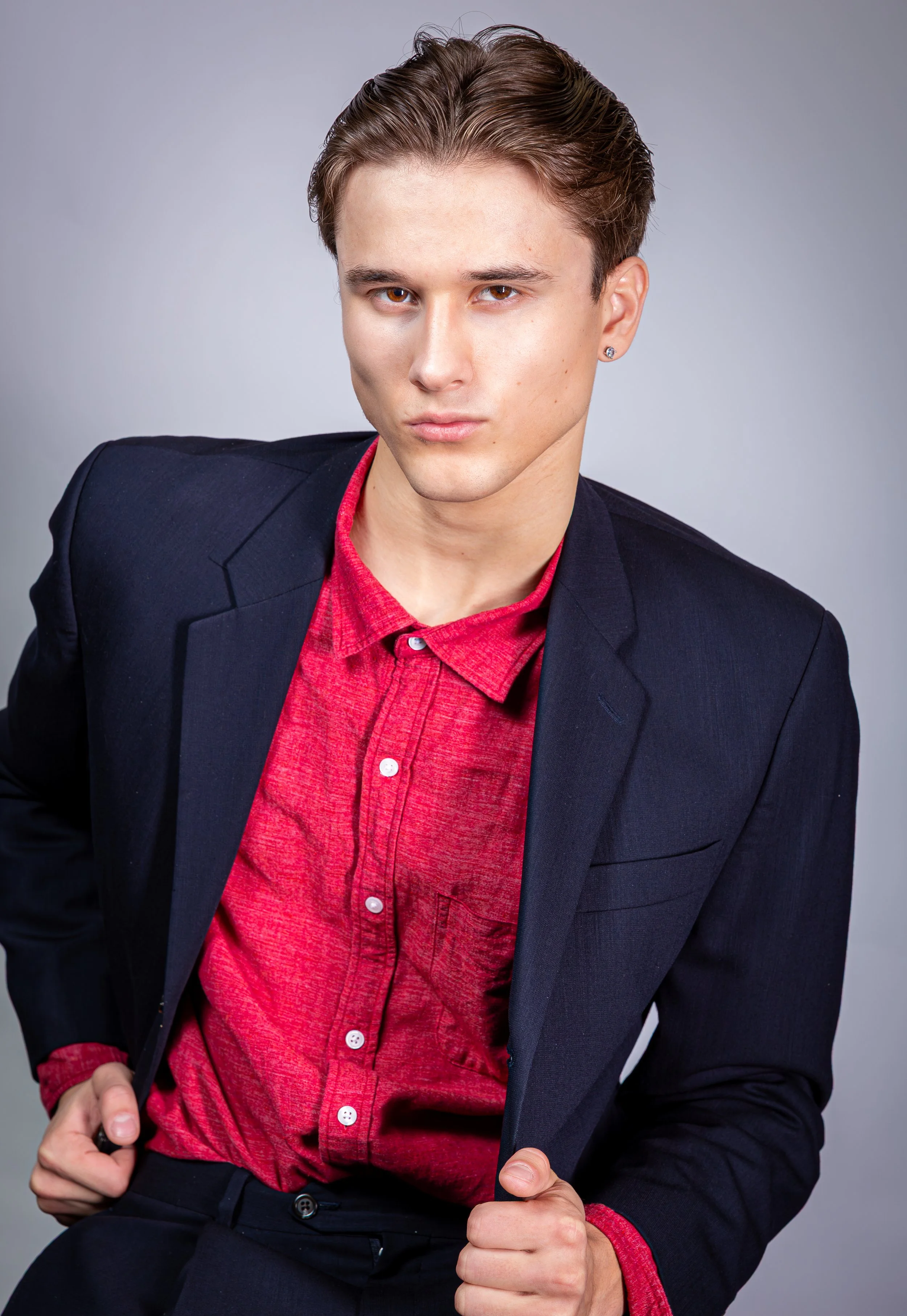 A young man with short brown hair wearing a black blazer over a red shirt, posing against a plain gray background.
