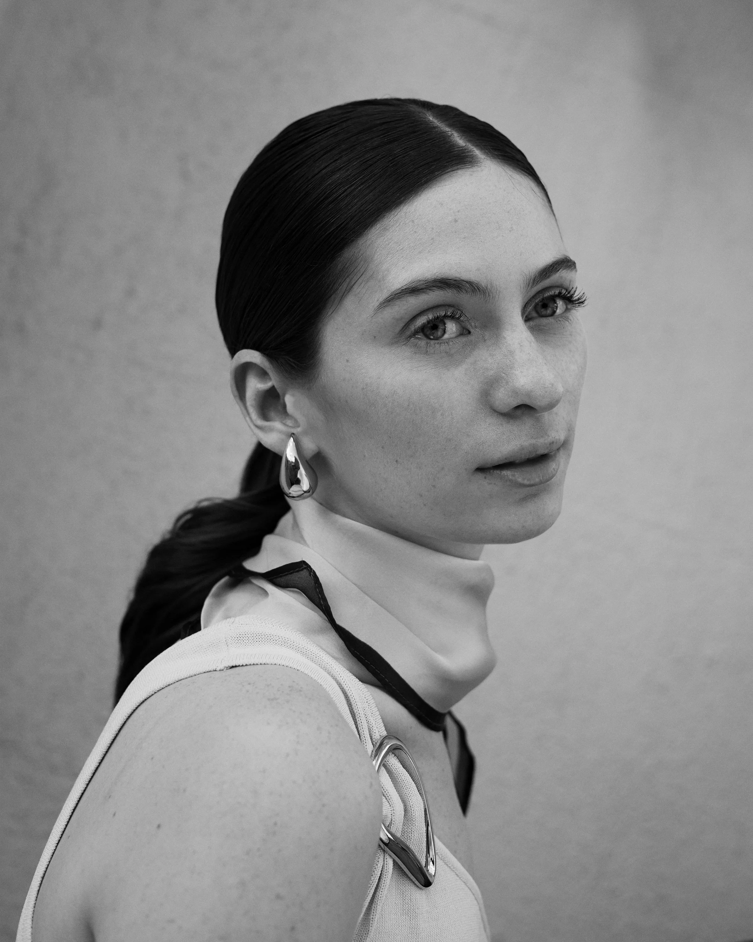 Black and white portrait of a woman with dark hair in a low ponytail, wearing a white sleeveless top, large teardrop-shaped earrings, and a high neck scarf, looking at the camera with a neutral expression.