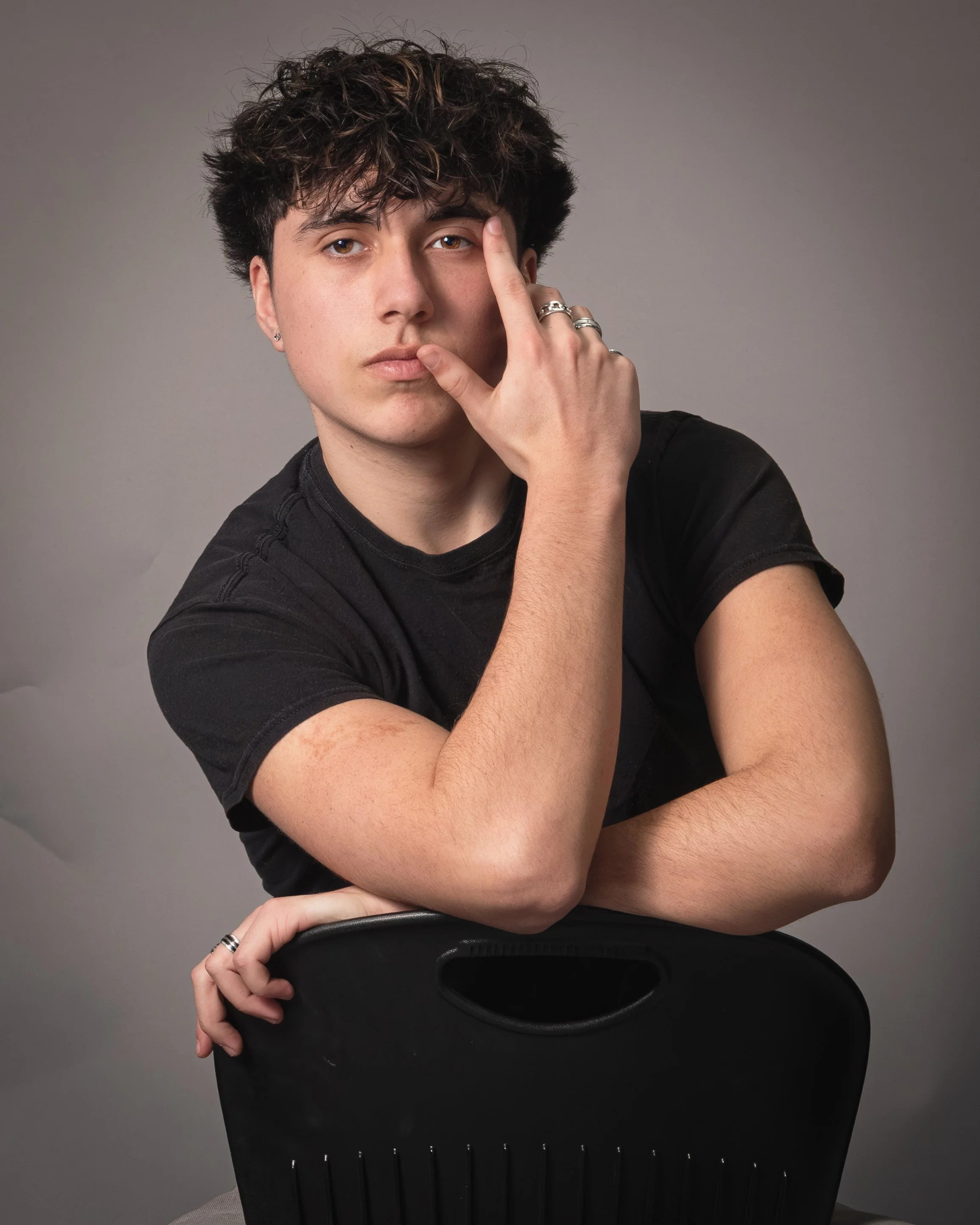 Young man with dark, curly hair and earrings, wearing a black t-shirt, holding a black chair with one arm and touching face with the other, against a plain gray background.