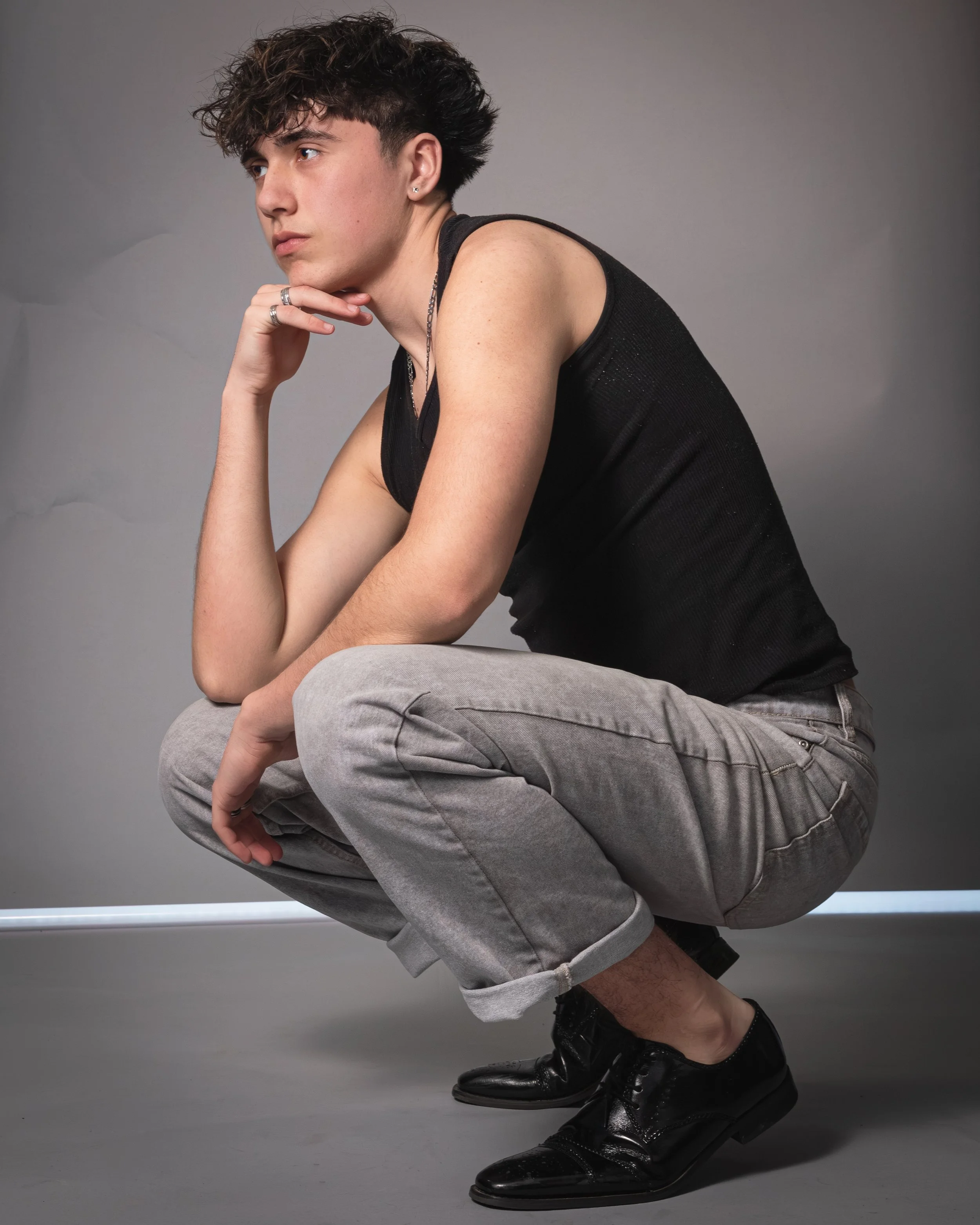 A young man with dark, curly hair squatting against a plain gray background, wearing a sleeveless black shirt, gray pants, and shiny black shoes, with his left hand resting near his chin.