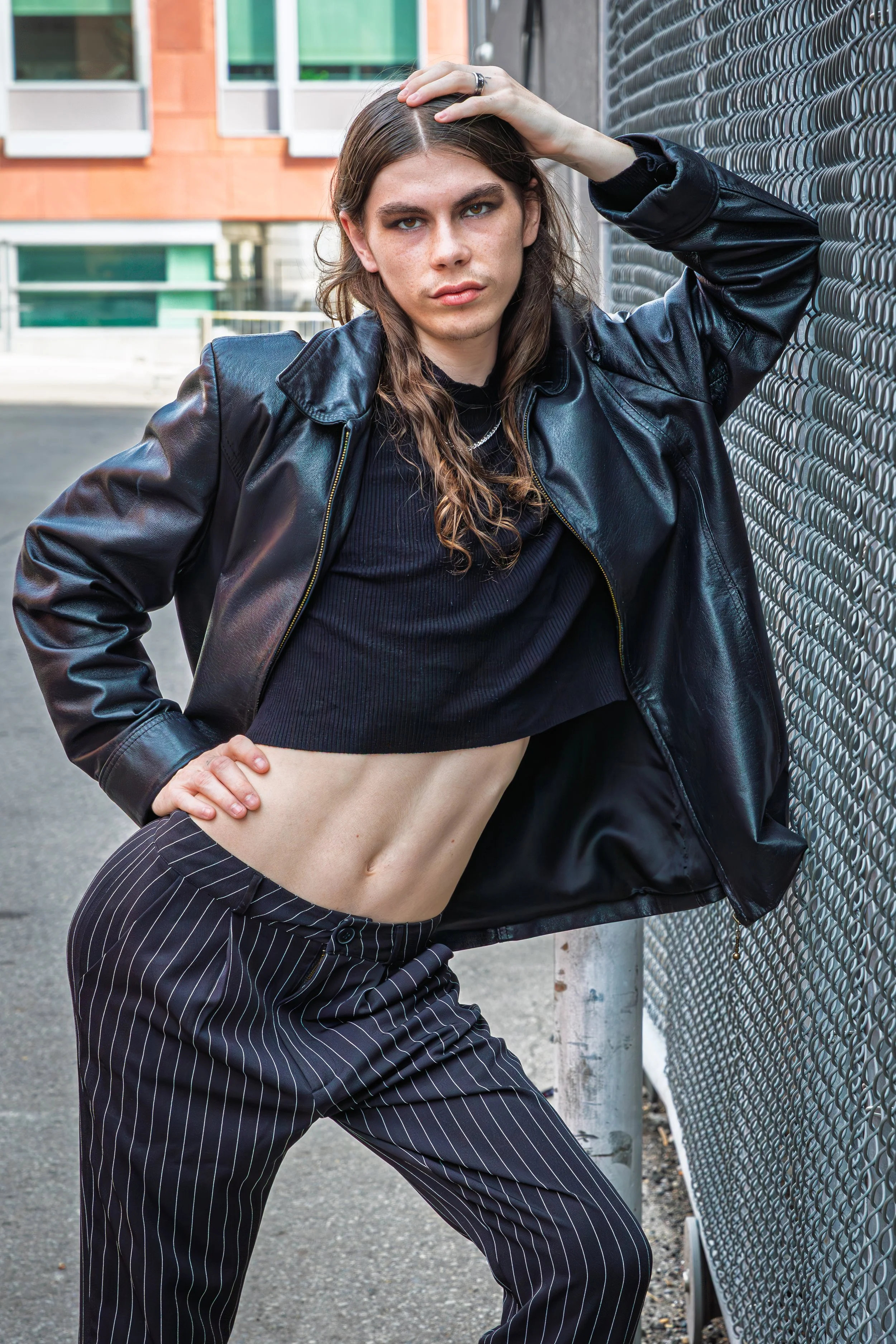 A young woman with long brown hair, wearing a black leather jacket, black crop top, and pinstriped pants, leans against a chain-link fence on an urban street.