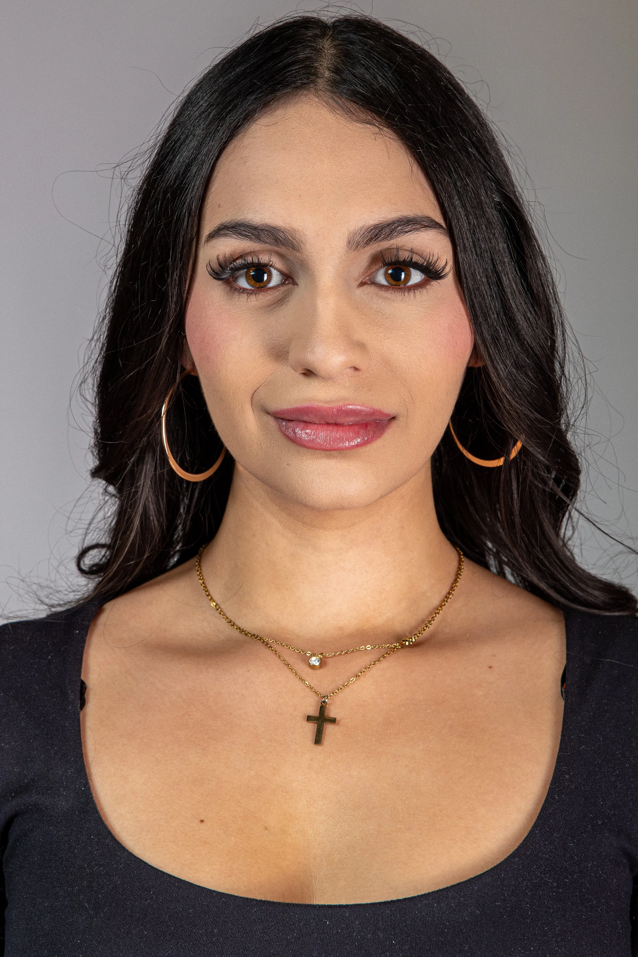 A young woman with long dark hair, wearing hoop earrings and layered gold necklaces with a cross pendant, smiling slightly against a gray background.