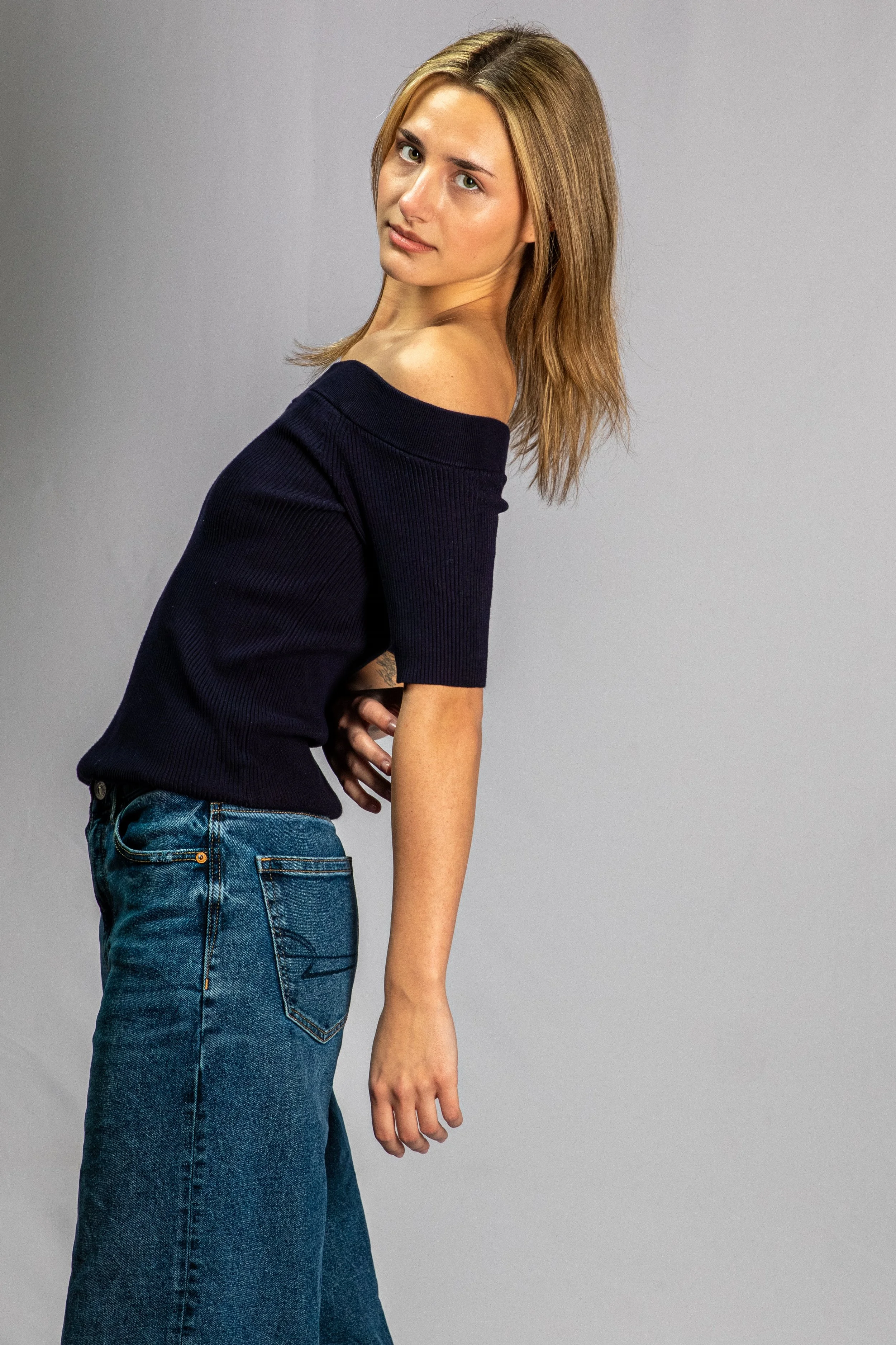 A young woman with shoulder-length light brown hair wearing a off-the-shoulder black top and blue jeans standing against a plain grey background, looking at the camera with a slight smile.