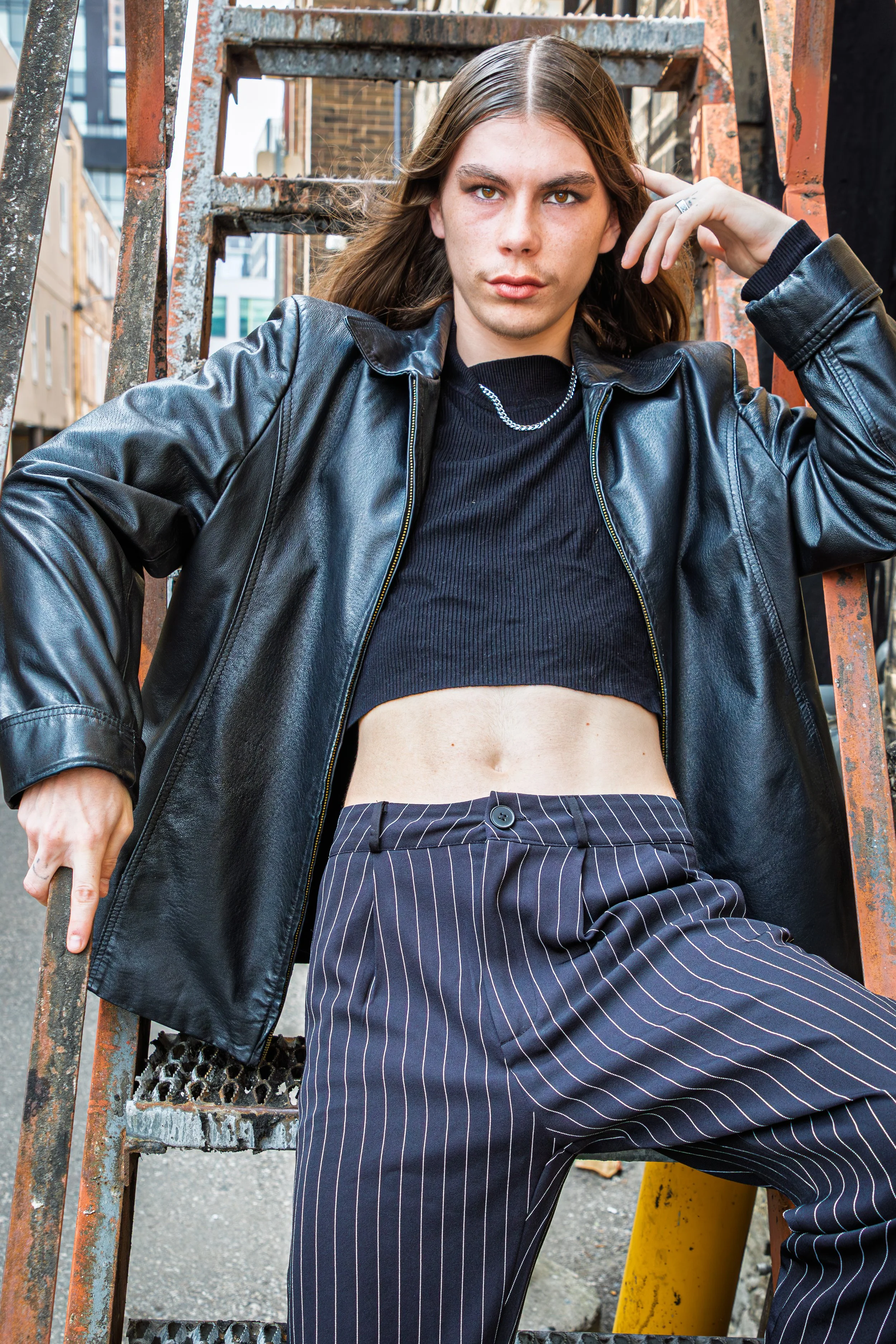 A young woman with long brown hair and defined eyebrows posing outdoors on a metal staircase, wearing a black leather jacket, crop top, and striped pinstripe pants.