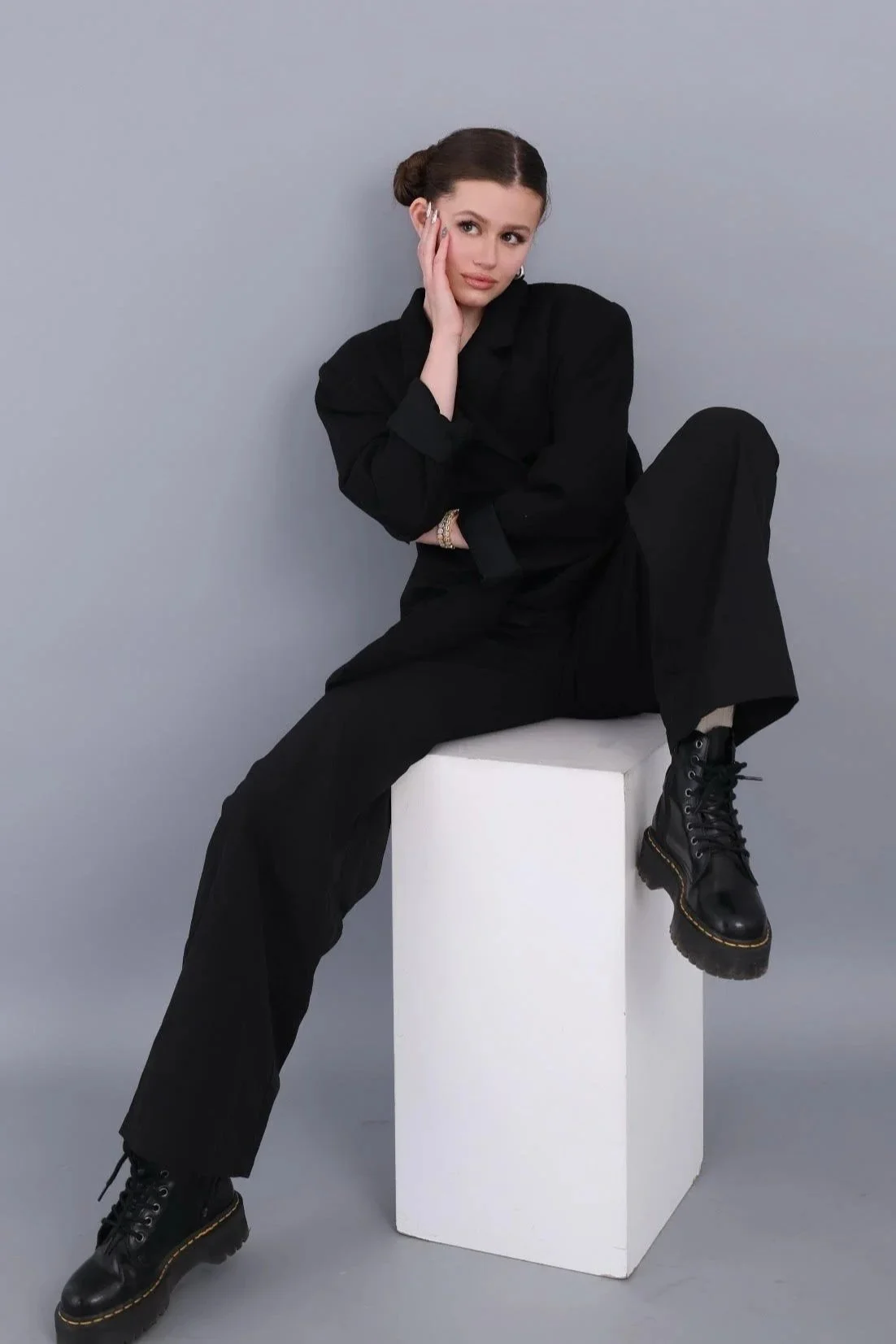 A woman with brown hair in buns, wearing a black outfit and boots, sitting on a white block against a gray background, resting her head on her hand, looking at the camera.