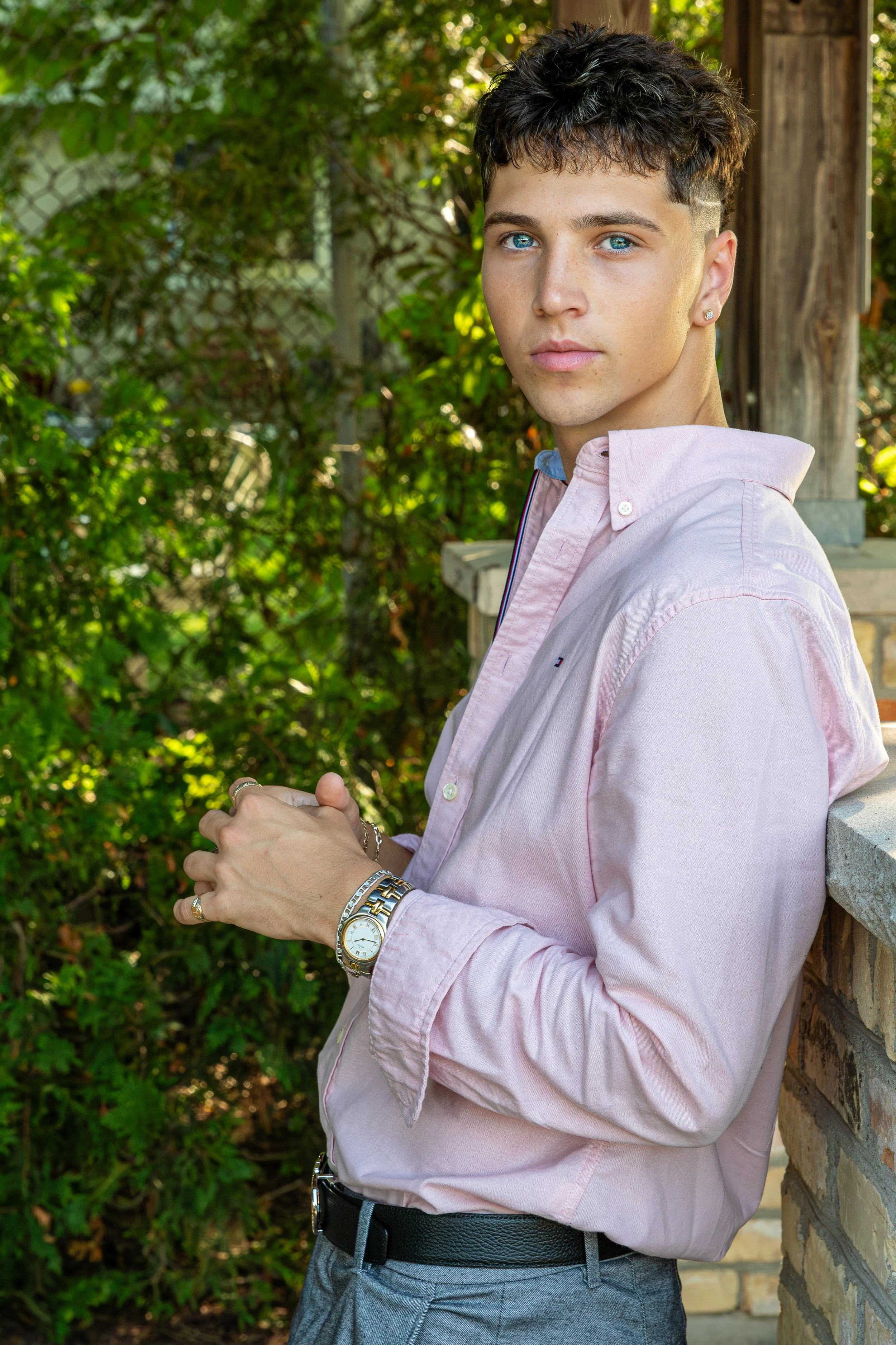 Young man with short dark curly hair and bright blue eyes, wearing a light pink button-up shirt with rolled sleeves, standing outdoors near a brick wall with greenery in the background.