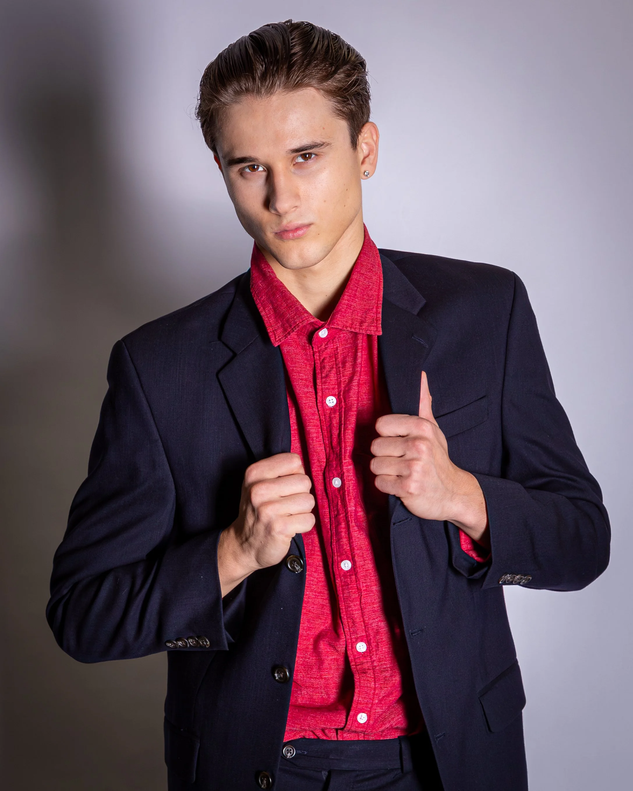 Young man in a dark suit and red shirt, holding the lapels of his suit jacket, looking directly at the camera against a plain light gray background.