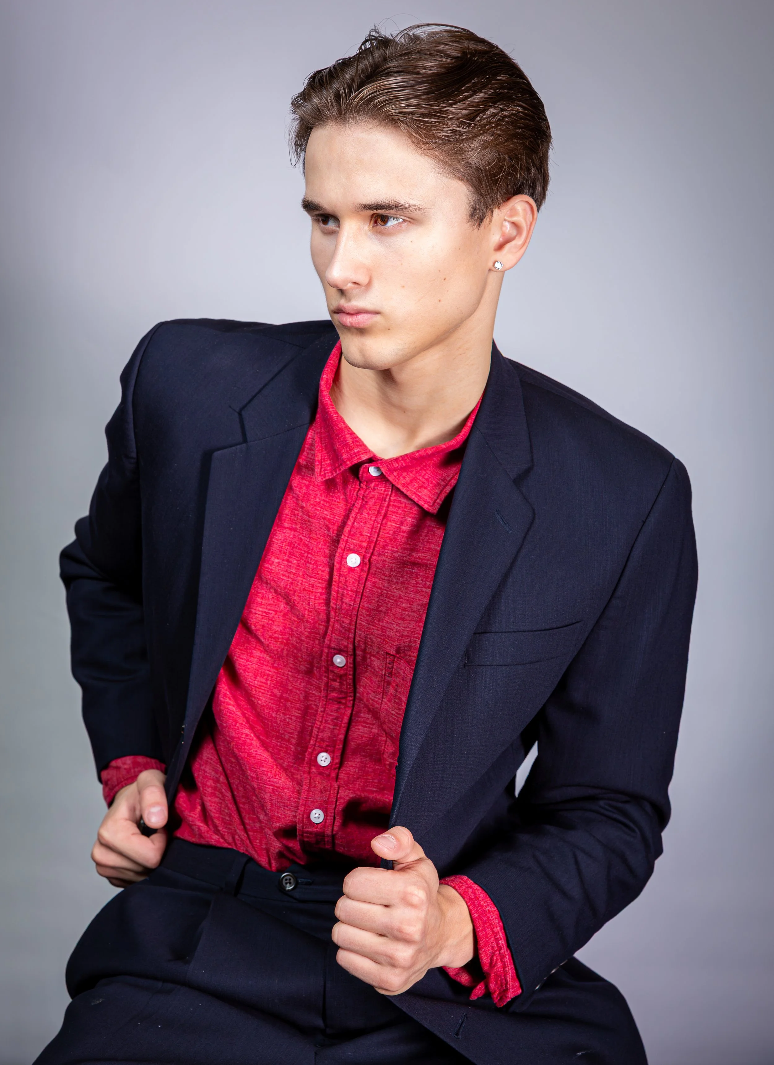 A young man with brown hair wearing a black blazer over a red shirt, sitting against a plain background.