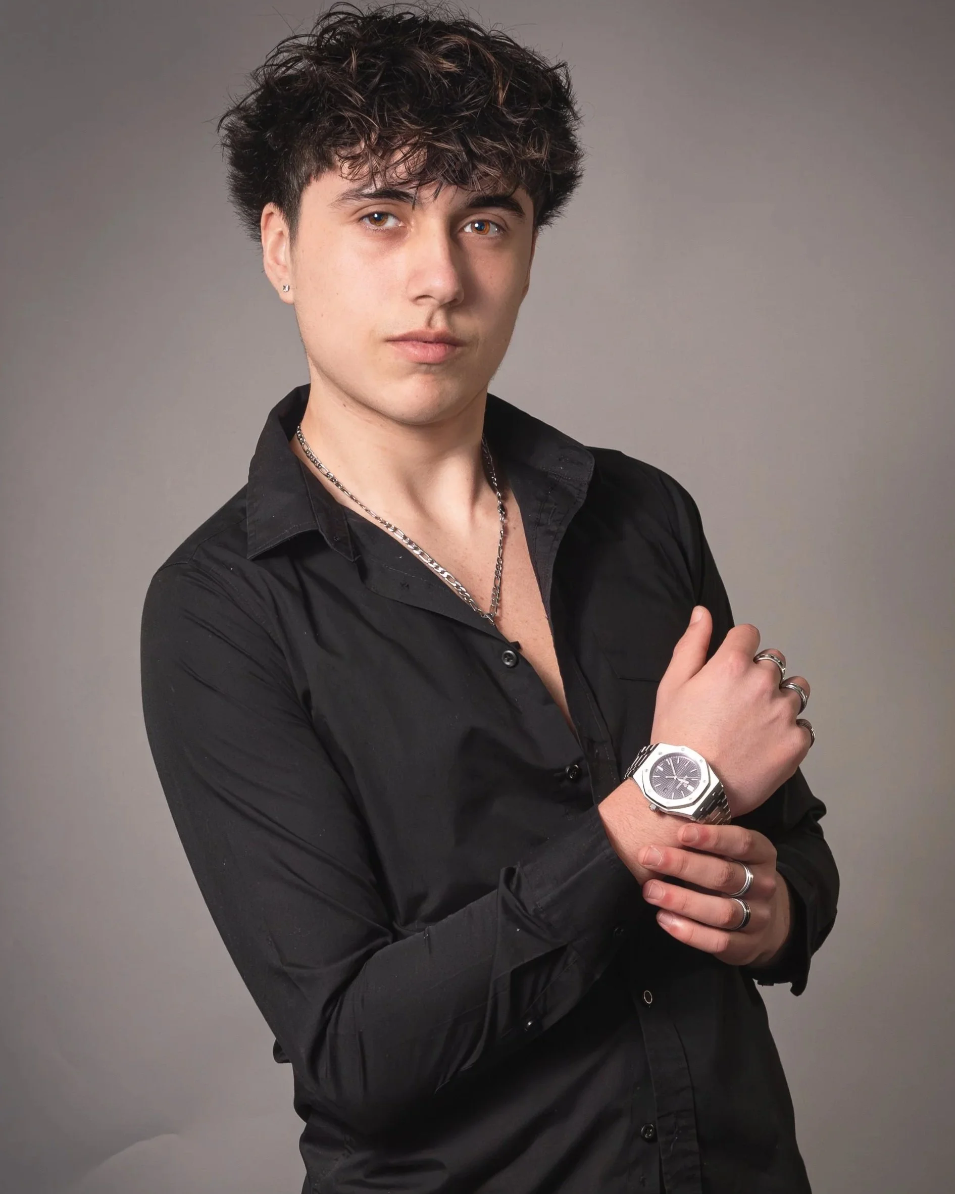 Young man with curly dark hair wearing a black button-up shirt, silver jewelry, and a wristwatch, posing against a gray background.