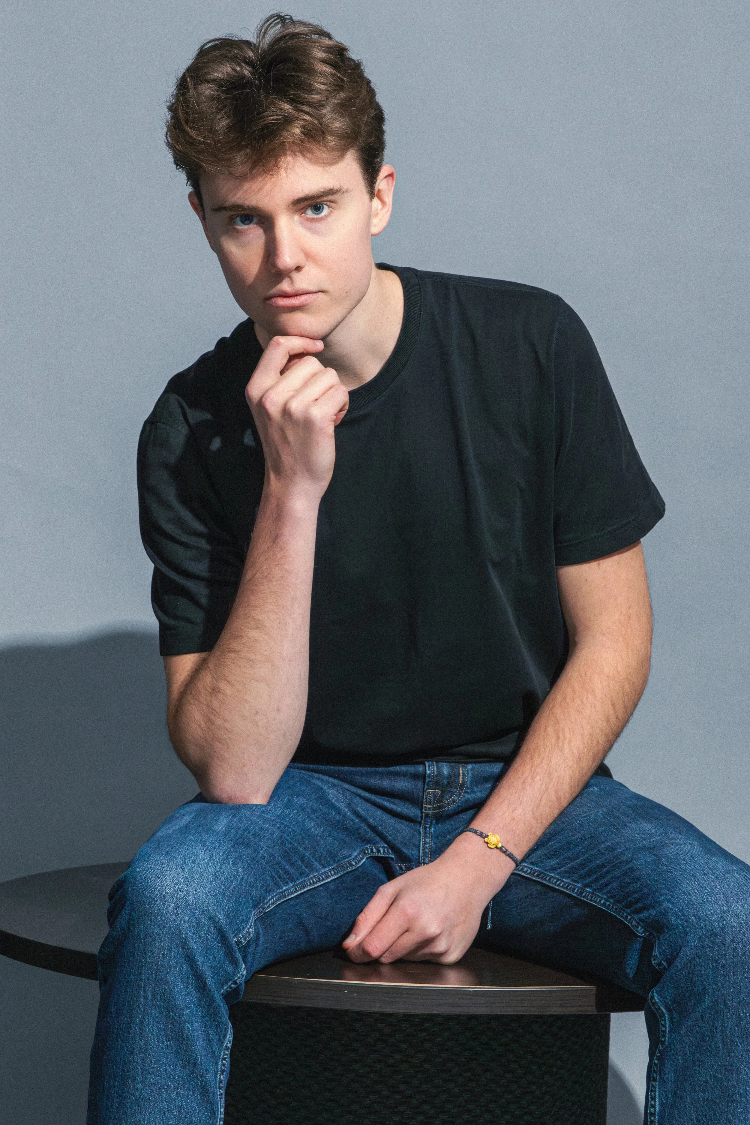 A young man with brown hair and blue eyes wearing a black t-shirt and blue jeans, seated on a black round stool, looking directly at the camera with his hand resting on his chin against a gray background.