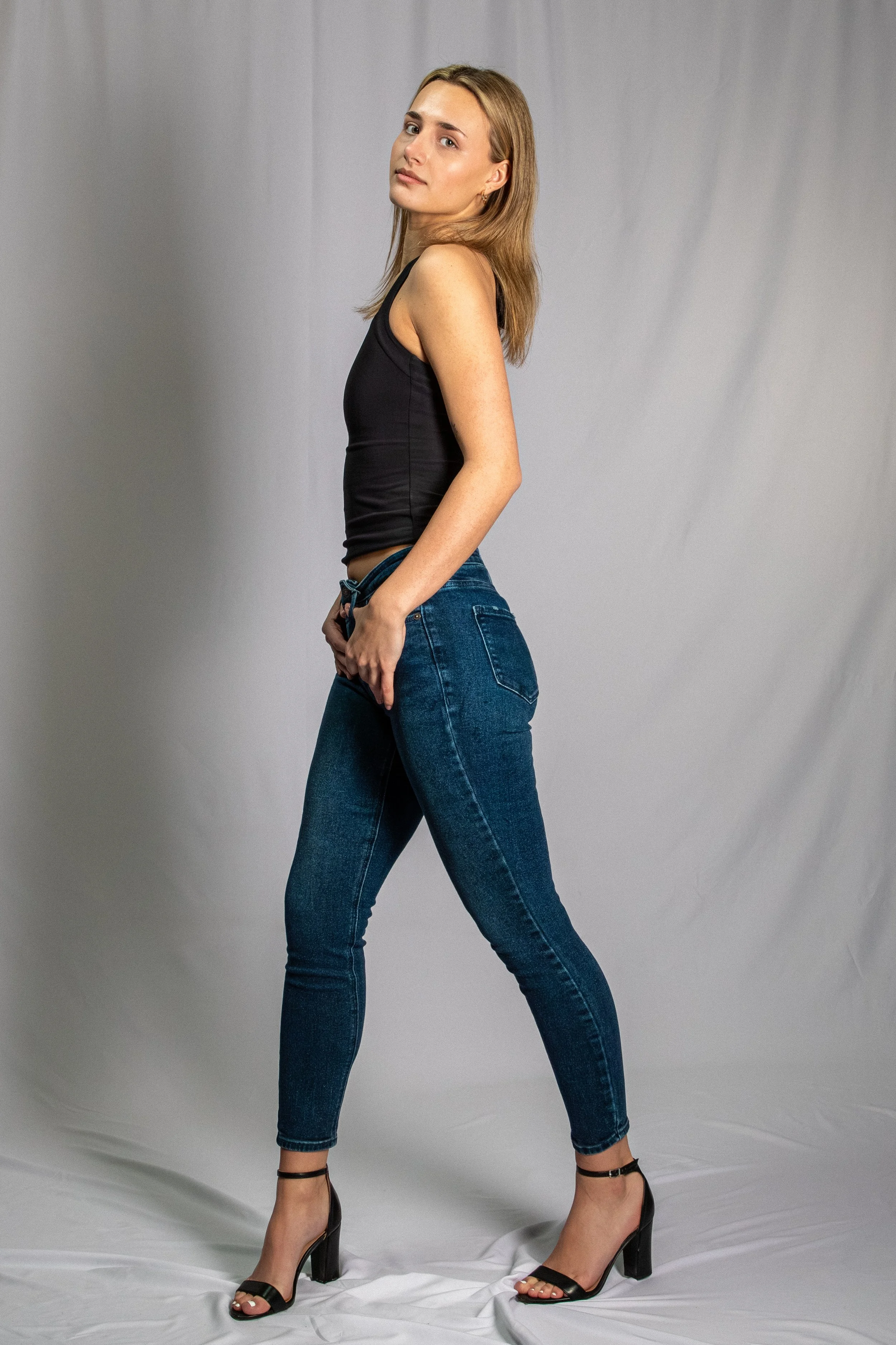 A young woman with blonde hair stands against a grey backdrop. She wears a black sleeveless top, blue jeans, and black high-heeled sandals. Her hand is resting on her hip and she is looking slightly to the side.