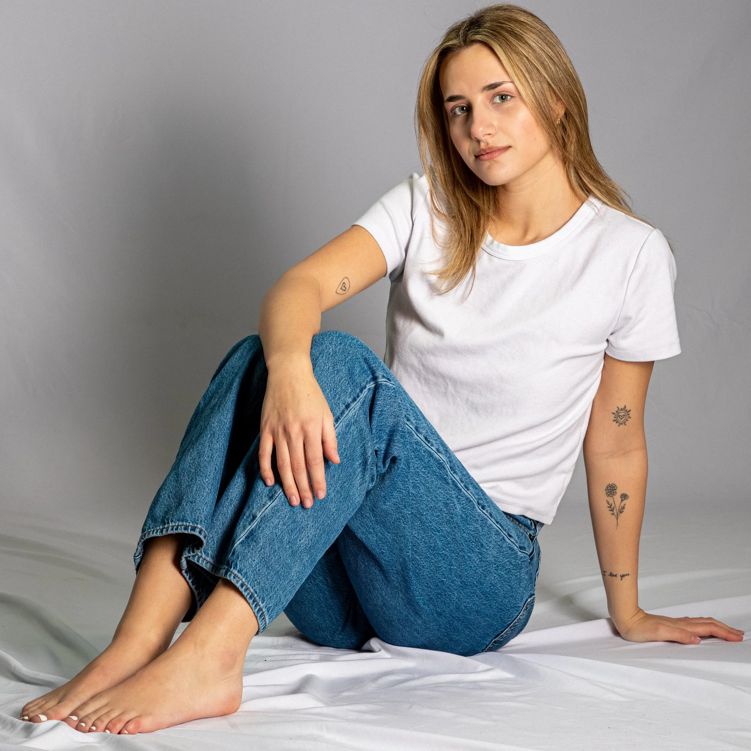 Woman sitting on the floor against a plain gray background, wearing a white t-shirt and blue jeans, with tattoos on her left arm and a small heart tattoo on her right forearm. She has light brown hair and is looking at the camera with a slight smile.