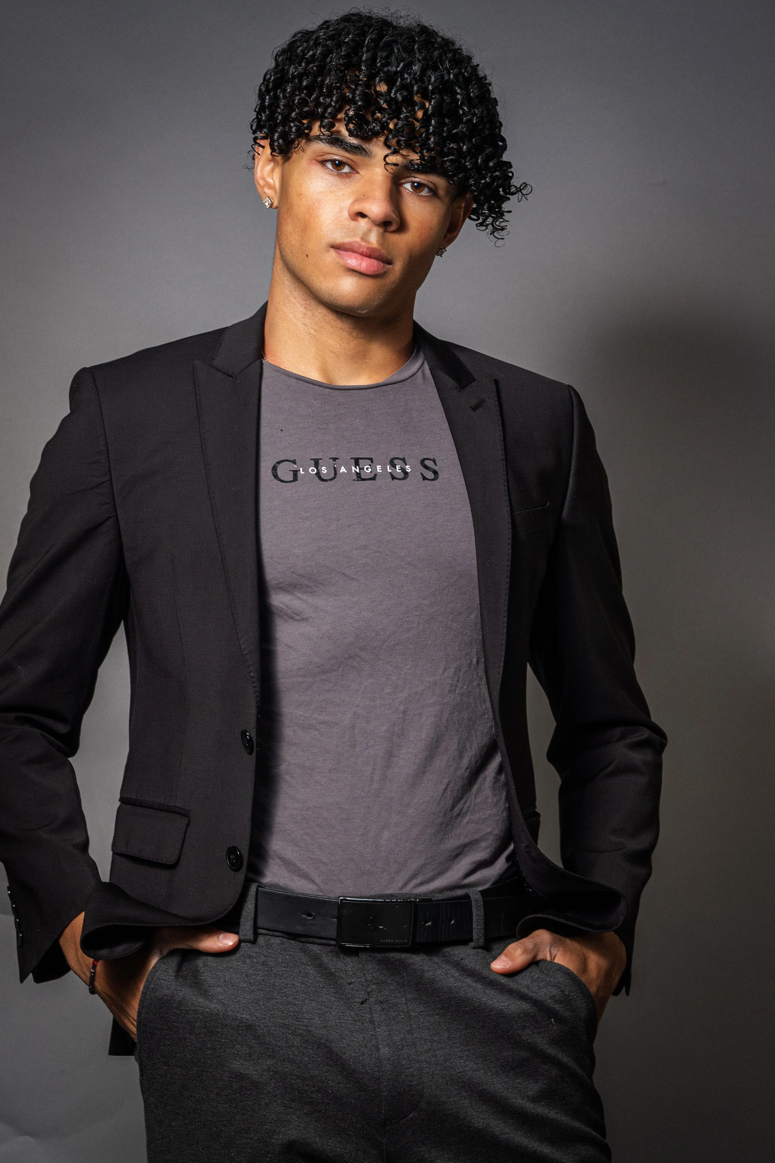 Young man with curly hair wearing a grey T-shirt with 'Guess' logo, black blazer, and grey pants, posing against a dark grey background.