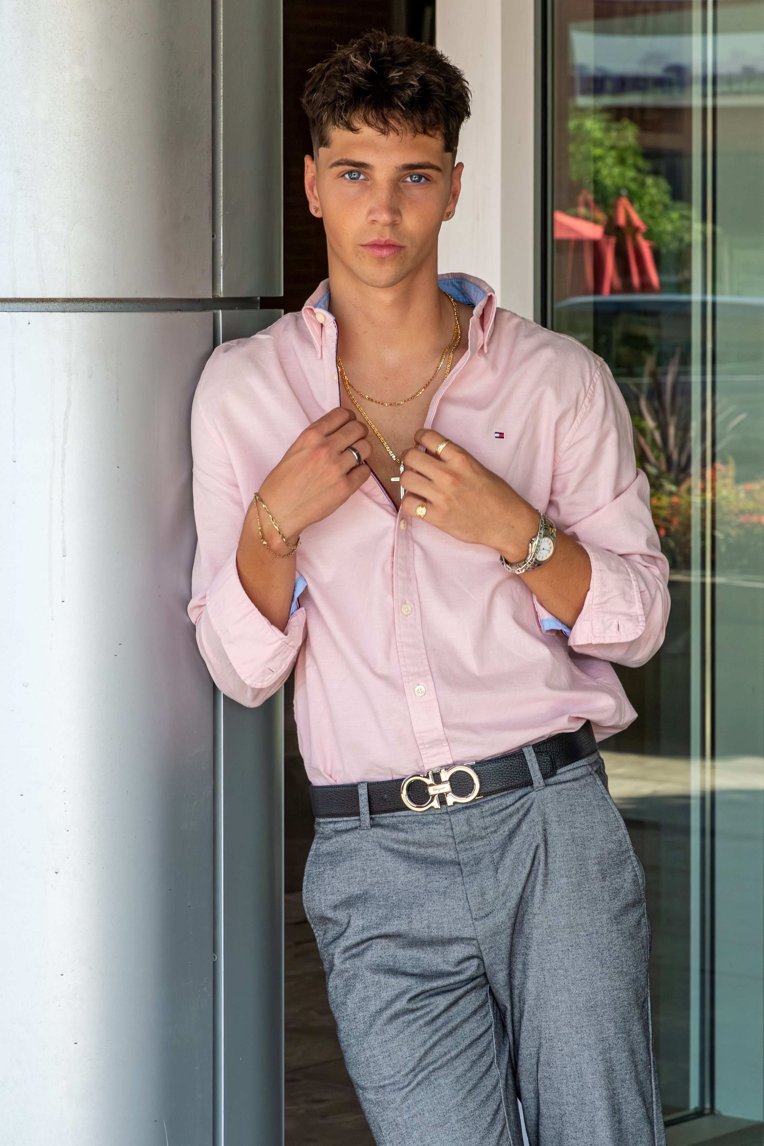 A young man with blue eyes and brown hair styled with a messy look, wearing a pale pink button-down shirt, gray trousers, and accessorized with gold jewelry including necklaces, rings, earrings, and a watch. He stands indoors near glass doors with a 