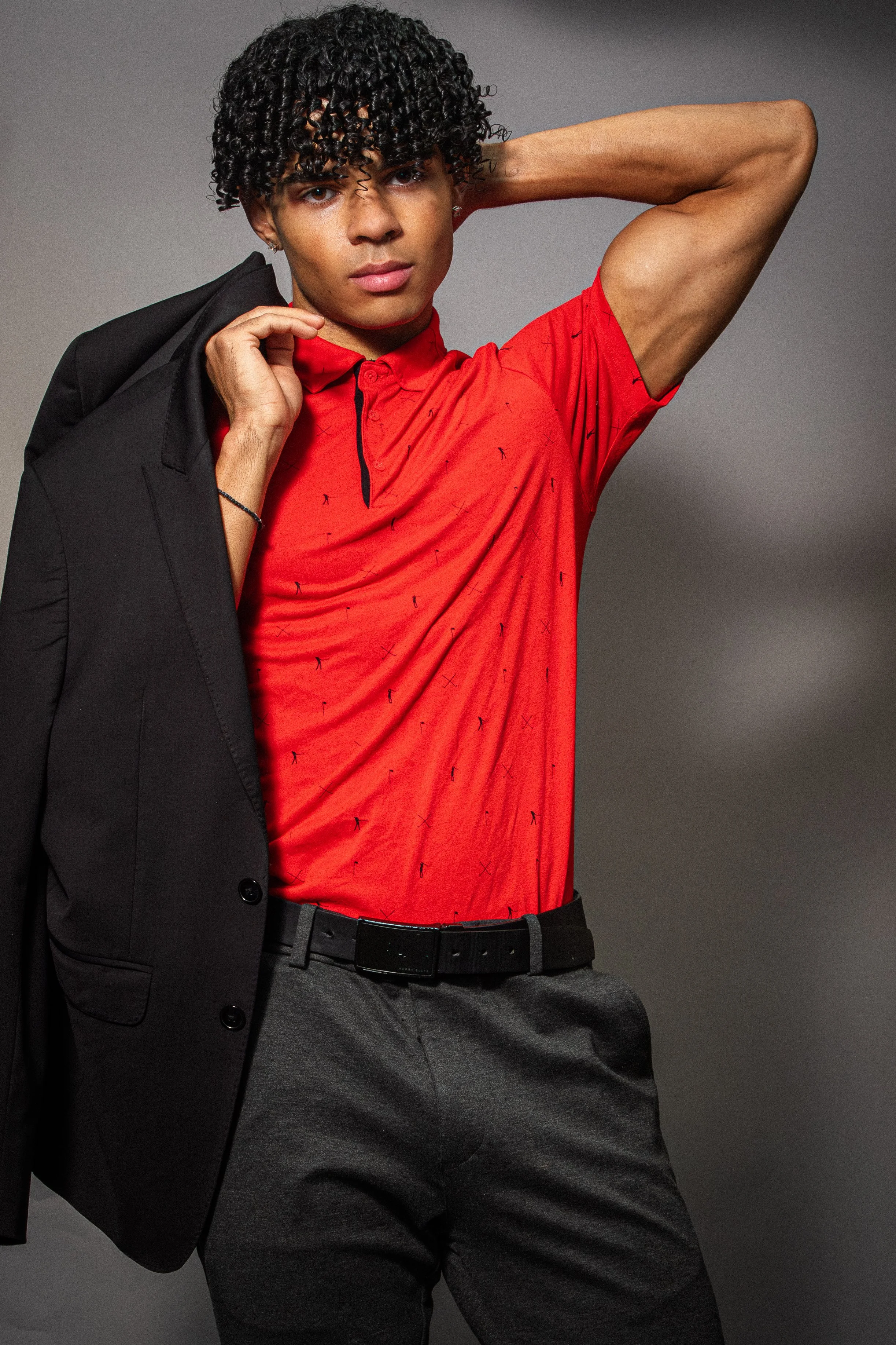 A young man with curly black hair and tan skin, wearing a red polo shirt with small black design and matching grey pants, holding a black blazer over his shoulder, posing against a plain grey background.