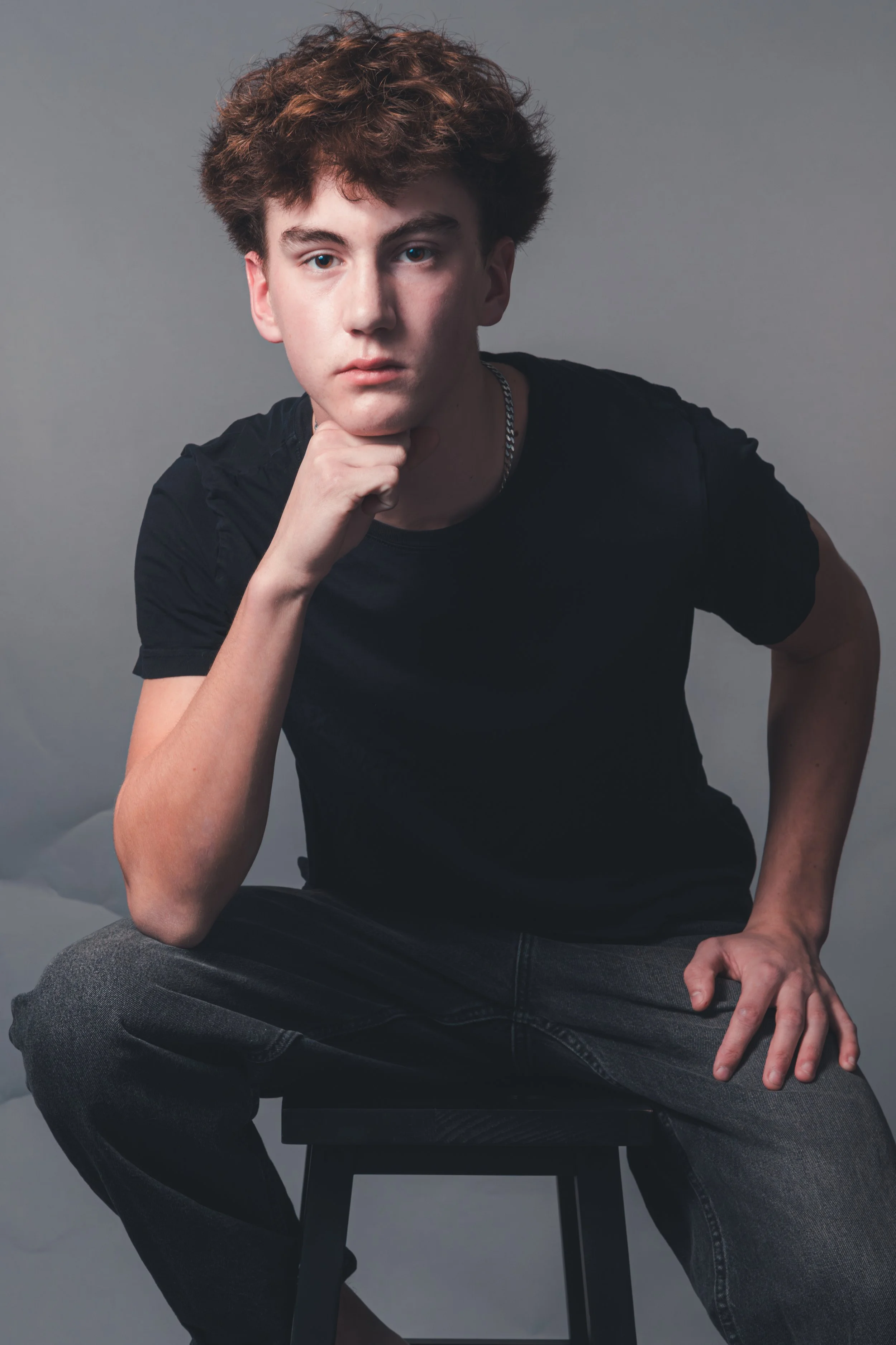 A young man with curly brown hair, dressed in a black T-shirt and jeans, sitting on a black stool against a gray background, resting his chin on his hand with a serious expression.