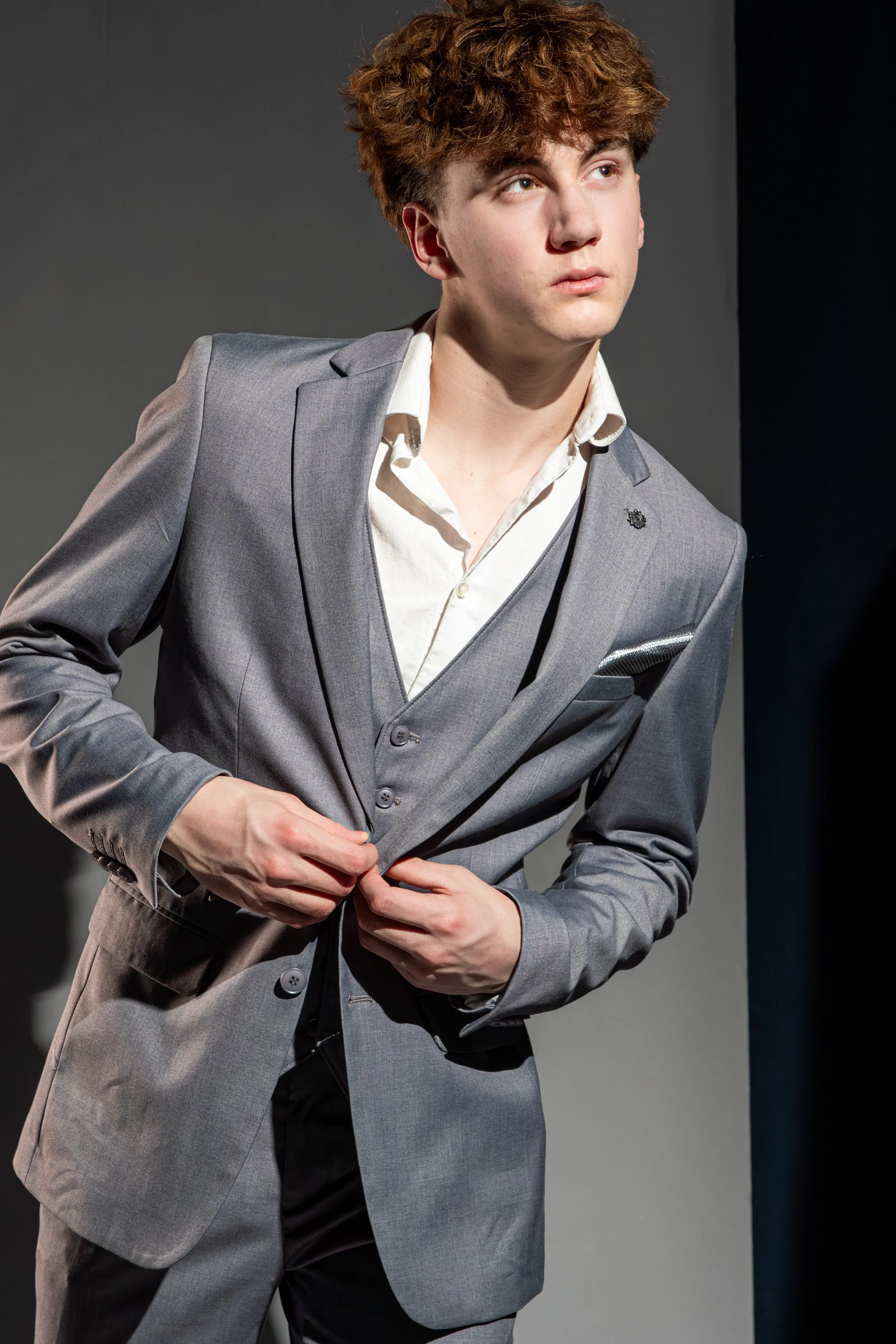 A young man with curly red hair wearing a gray suit and white shirt, adjusting his suit jacket while looking to the side against a plain dark background.