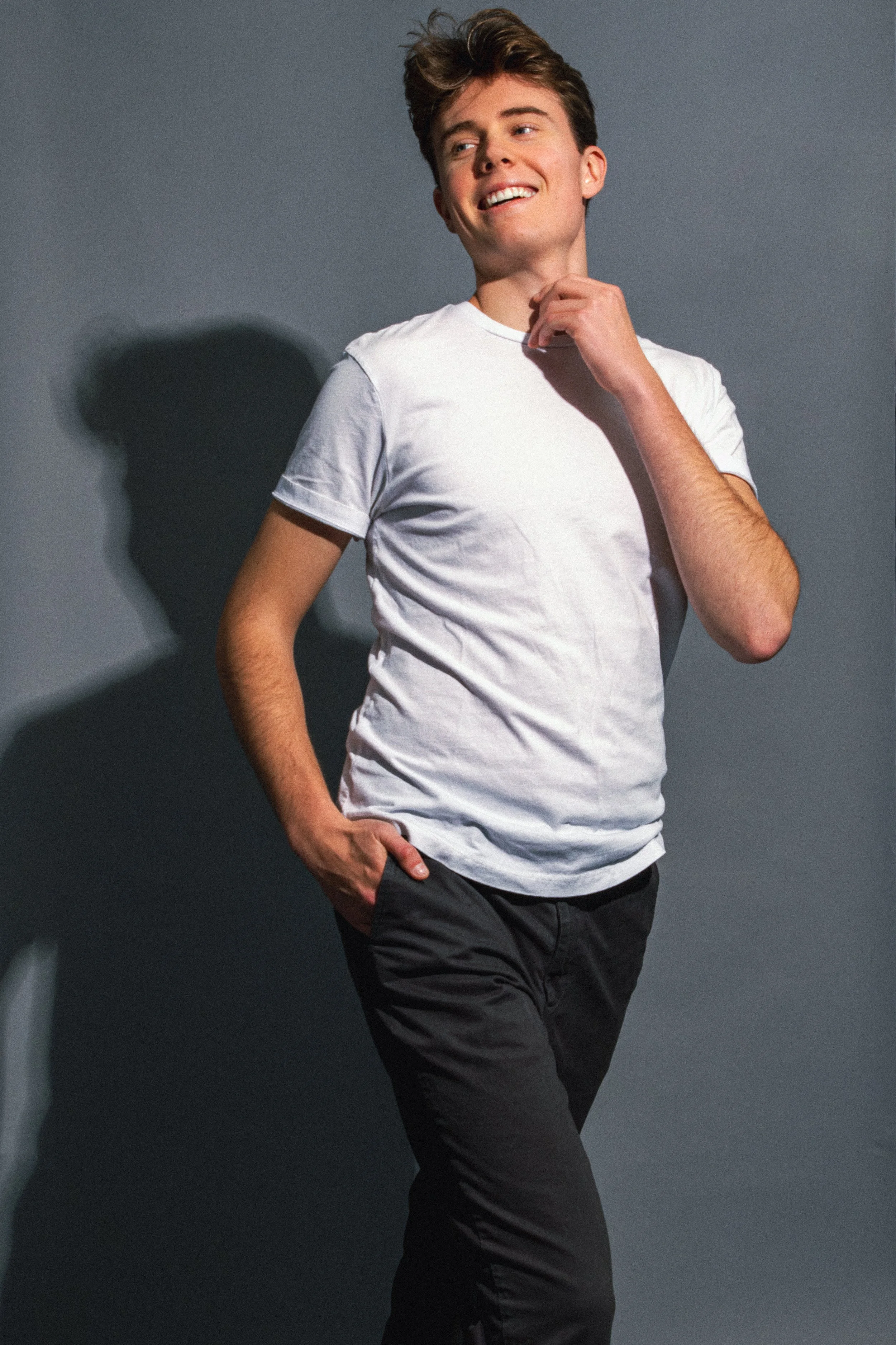 Young man in white t-shirt and black pants smiling, standing against gray background with shadow.