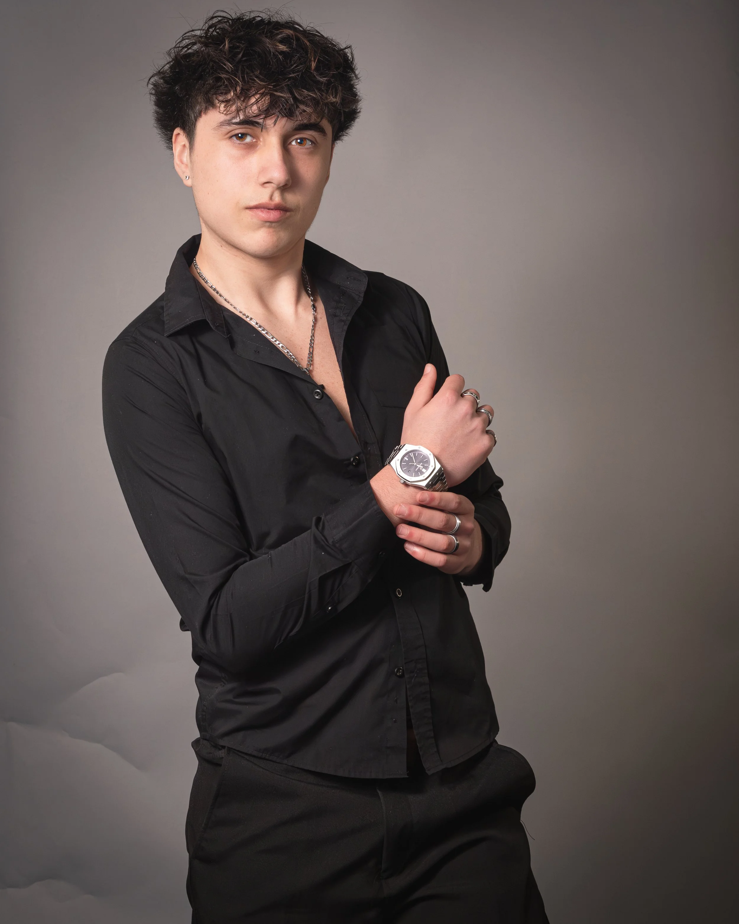 Young man with curly hair wearing a black button-up shirt, silver watch, and rings, posing with crossed arms against a gray background.