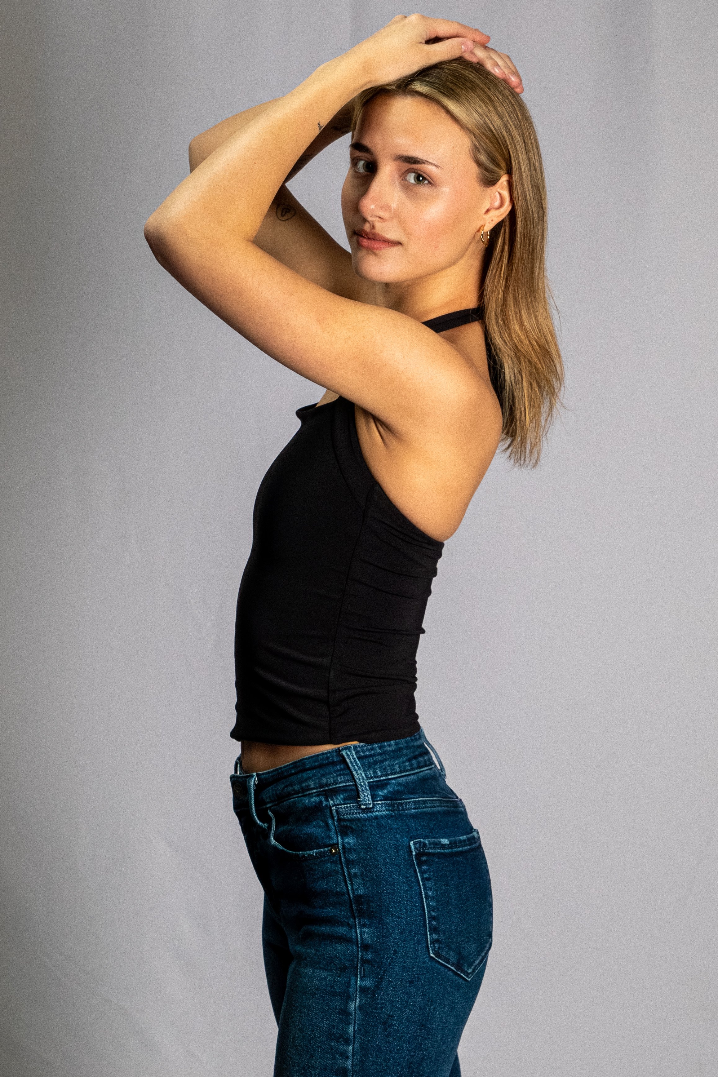 A young woman with light skin and shoulder-length light brown hair is posing with two hands resting on her head, wearing a black tank top and blue jeans, looking at the camera against a gray background.