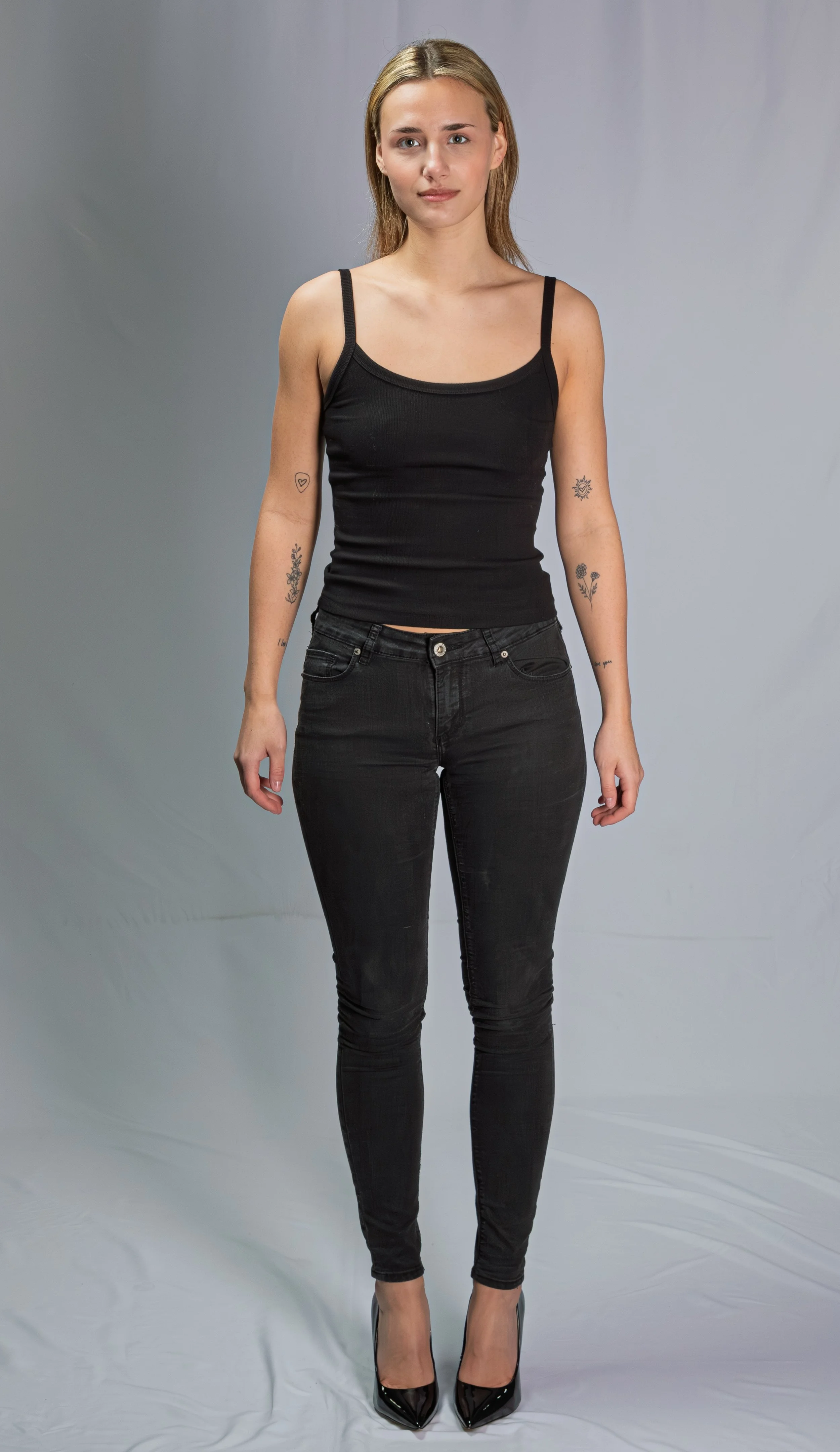 A young woman with long blond hair, wearing a black tank top, black jeans, and black high heels, standing against a plain background.