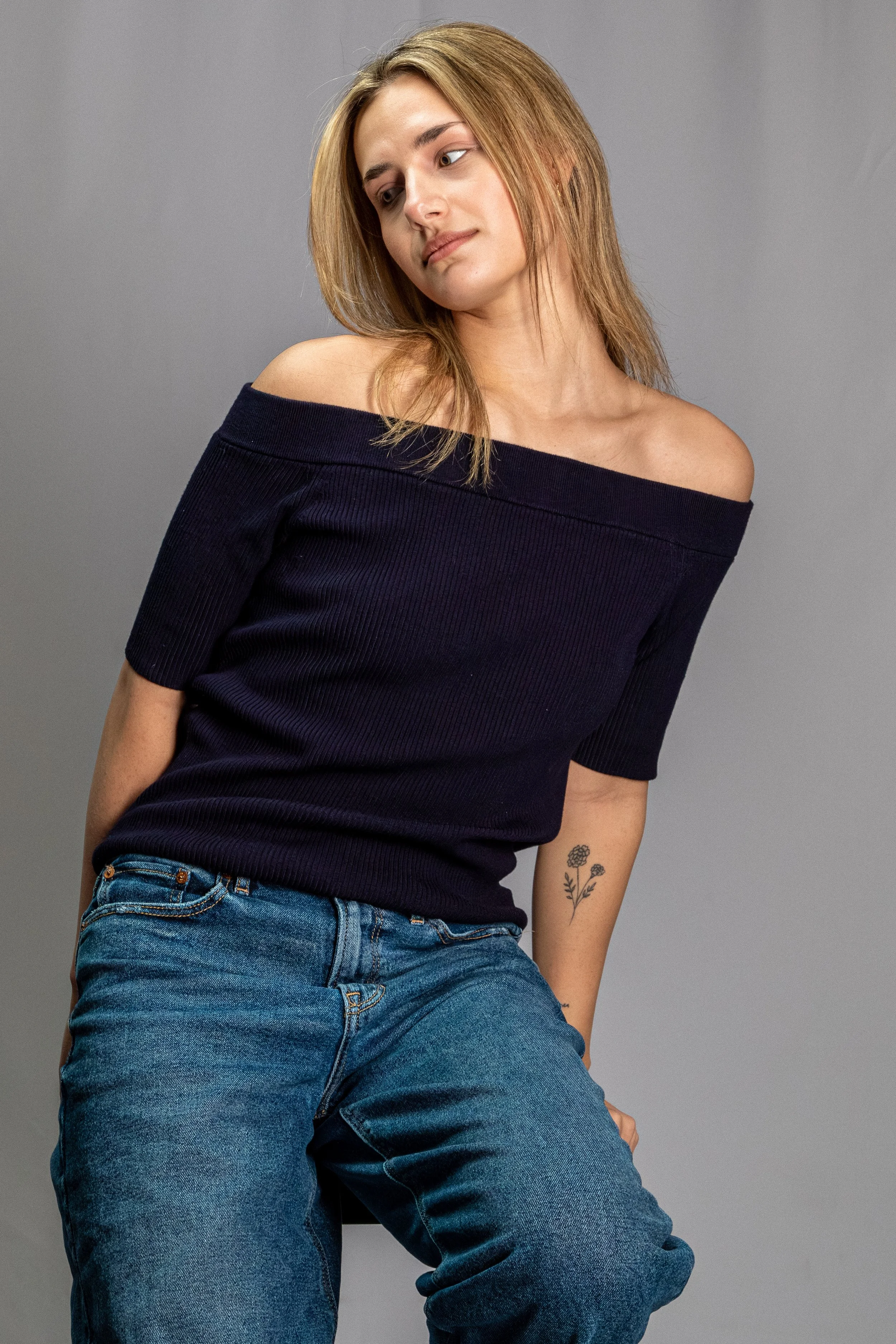 A woman with light skin and shoulder-length, blonde hair styled straight, posing against a plain gray background. She is wearing an off-the-shoulder black top and blue jeans, with a floral tattoo on her left forearm, looking downward with a gentle ex