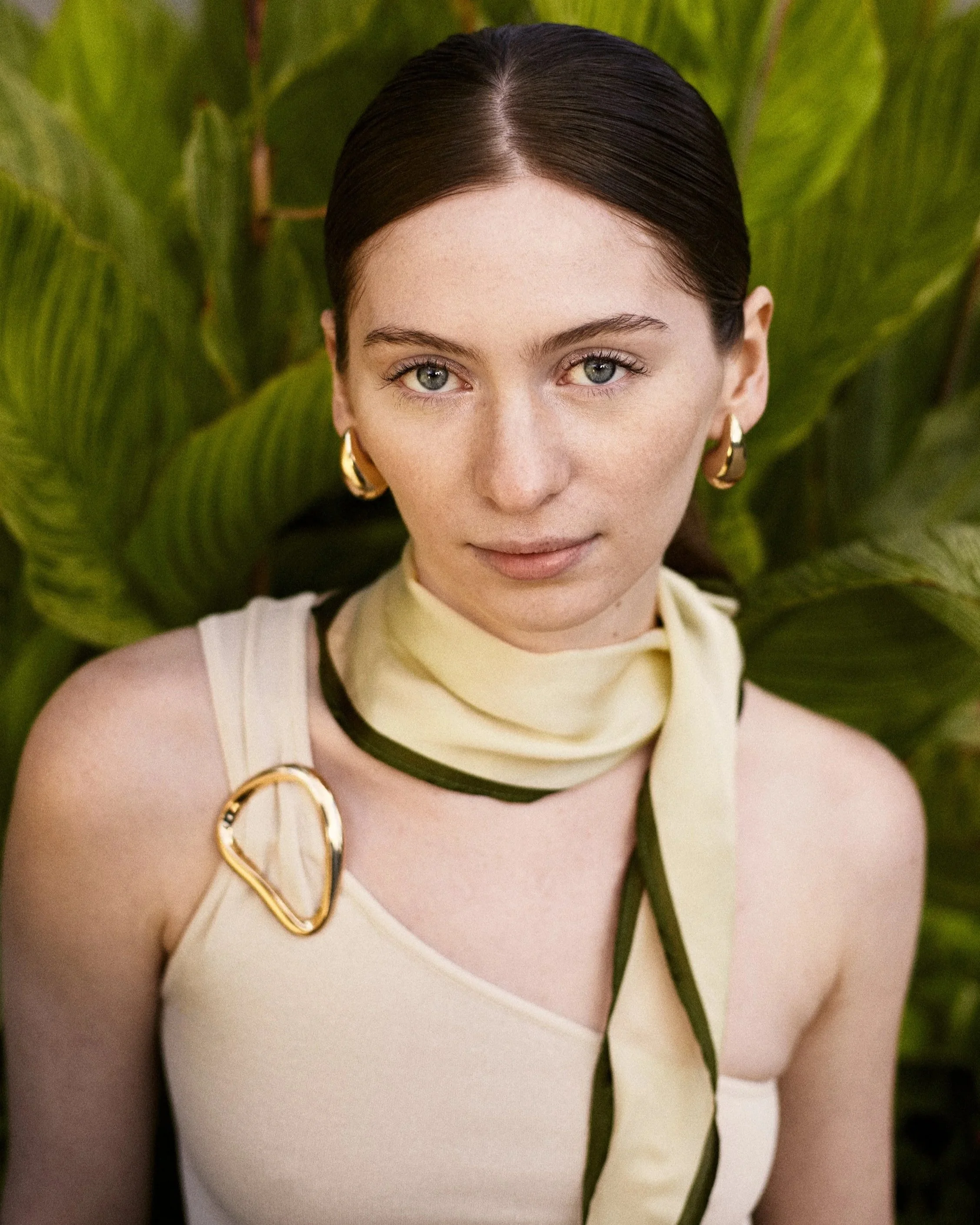 Close-up portrait of a woman with dark hair, blue eyes, and gold hoop earrings, wearing a cream top with black accents and a cream and green scarf, standing in front of large green tropical leaves.