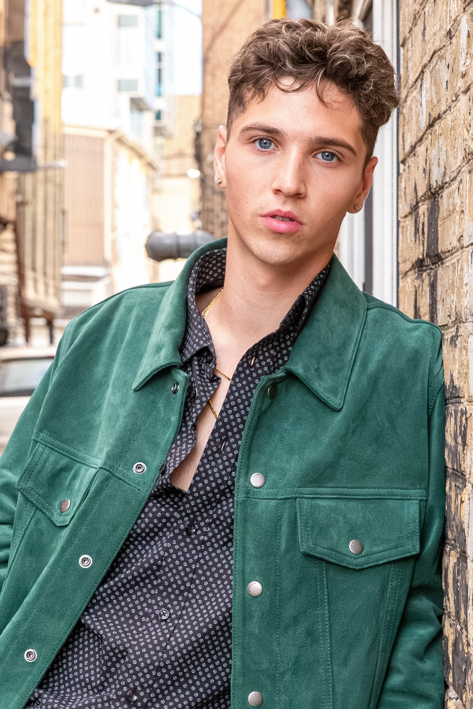 A young person with short, curly brown hair and blue eyes leaning against a brick wall in an urban alleyway, wearing a green jacket over a black button-up shirt with a white pattern.