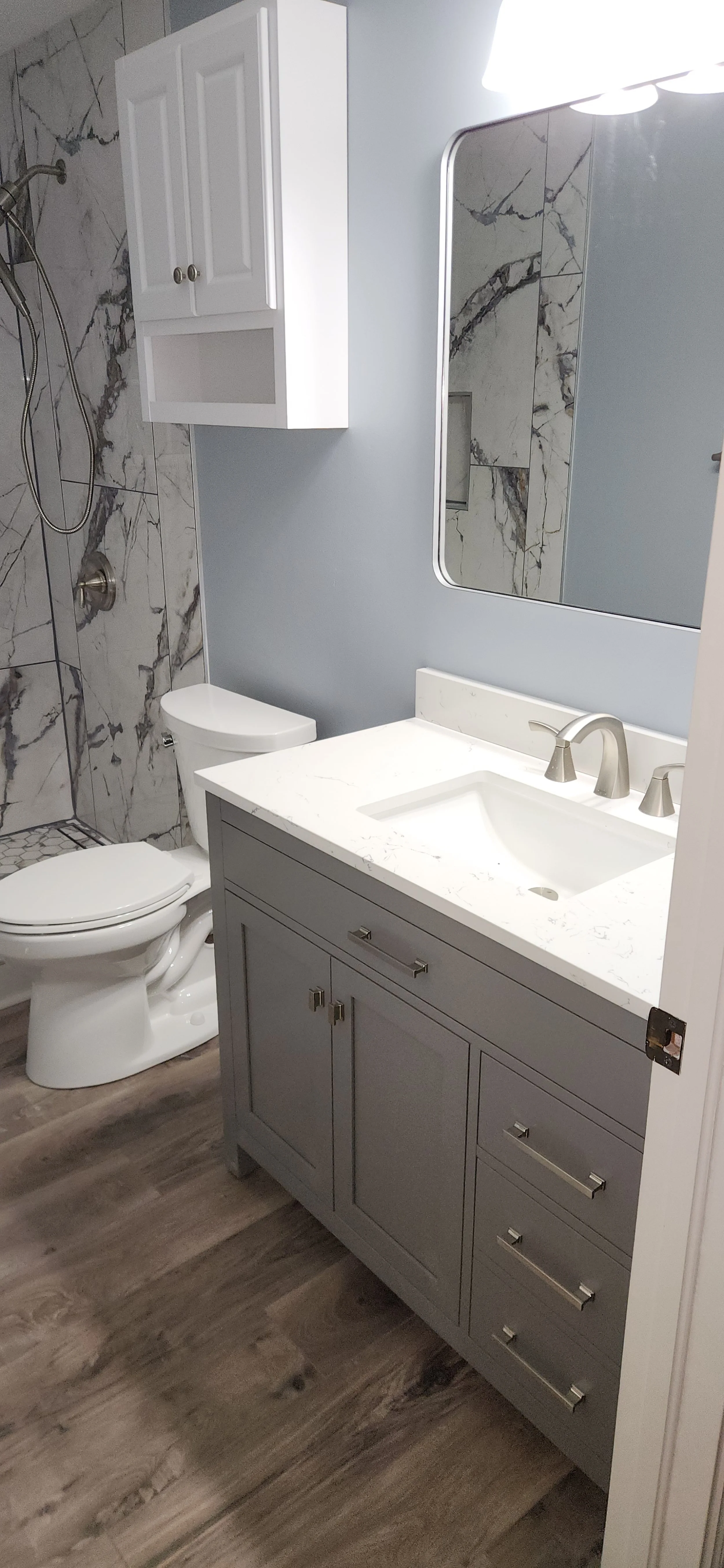 Bathroom with gray vanity, white marble countertop, and a mirror. There is a toilet and a wall cabinet in the background. The shower area has marble tiles and a showerhead.