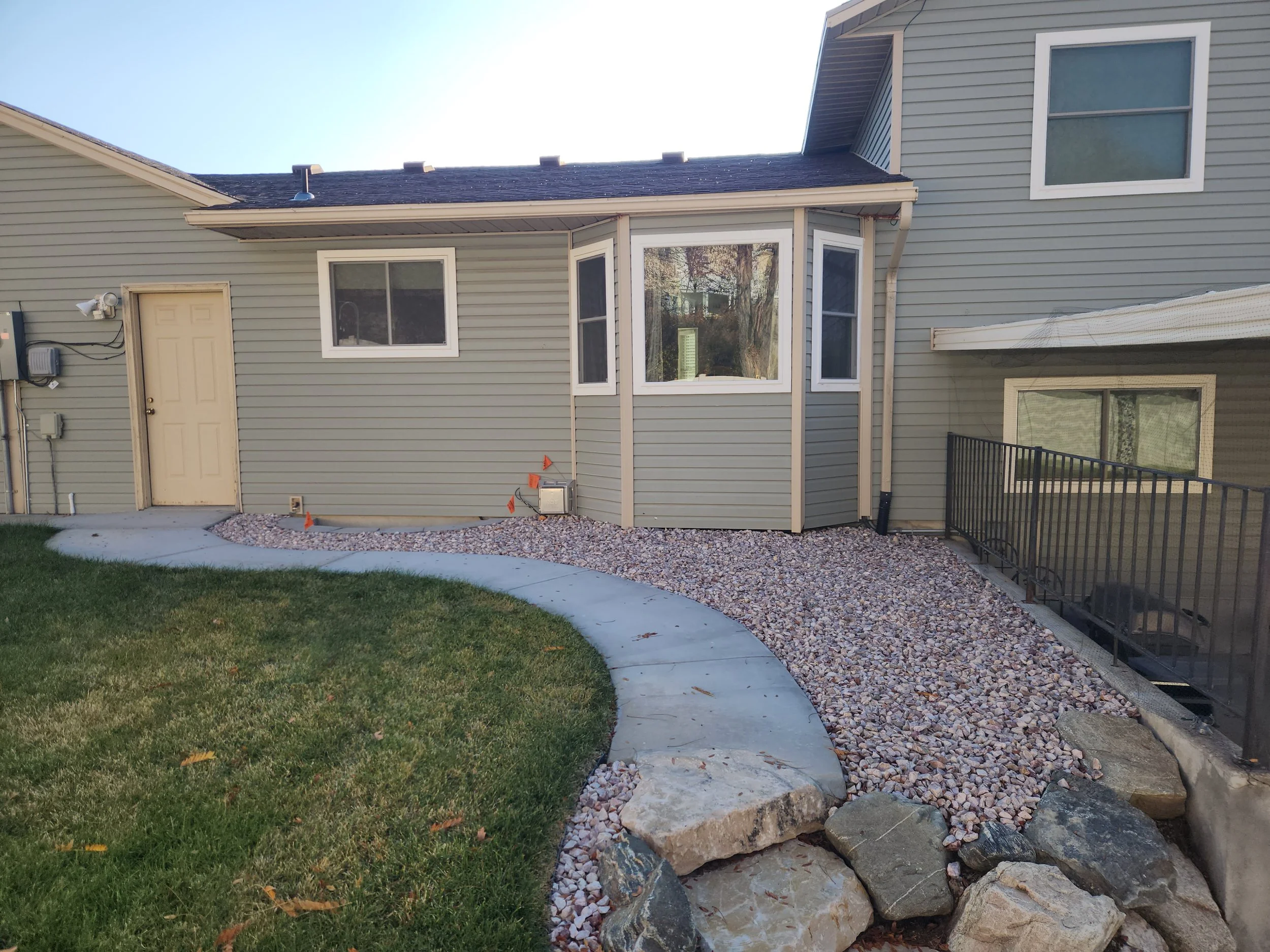 Backyard with a curved concrete pathway bordered by a grassy area and decorative rocks leading to a house with gray siding and multiple windows.