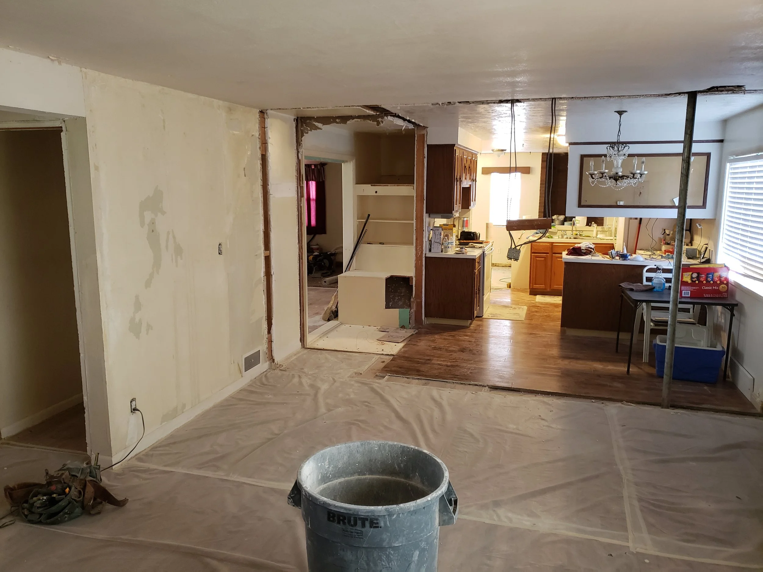 Interior of a living room and kitchen under renovation, with torn-down wall, construction materials, and plastic covering on the floor.