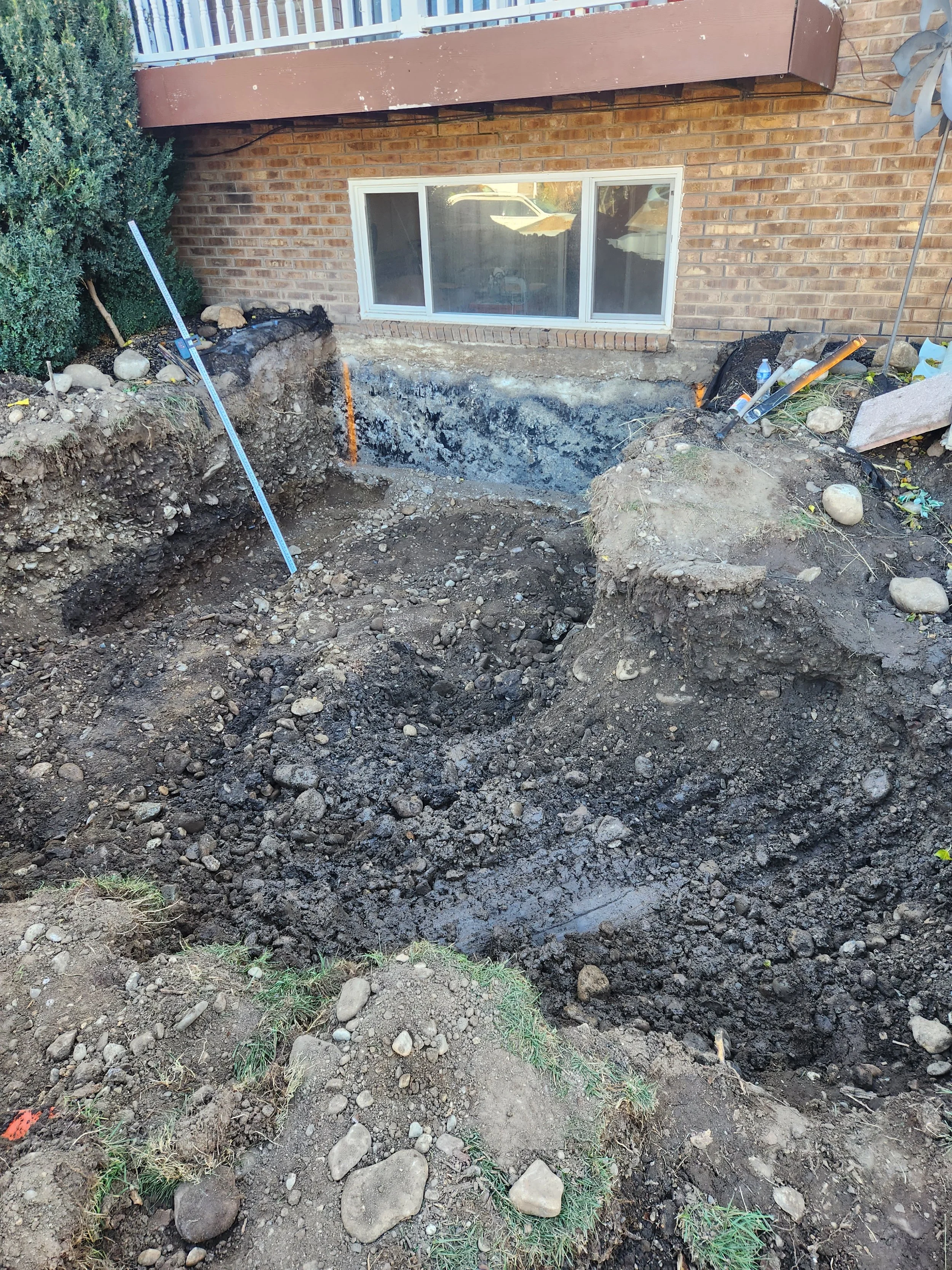 Construction site with dug-out soil in front of a brick house, showing an excavation for foundation or utility work, with tools and equipment nearby.